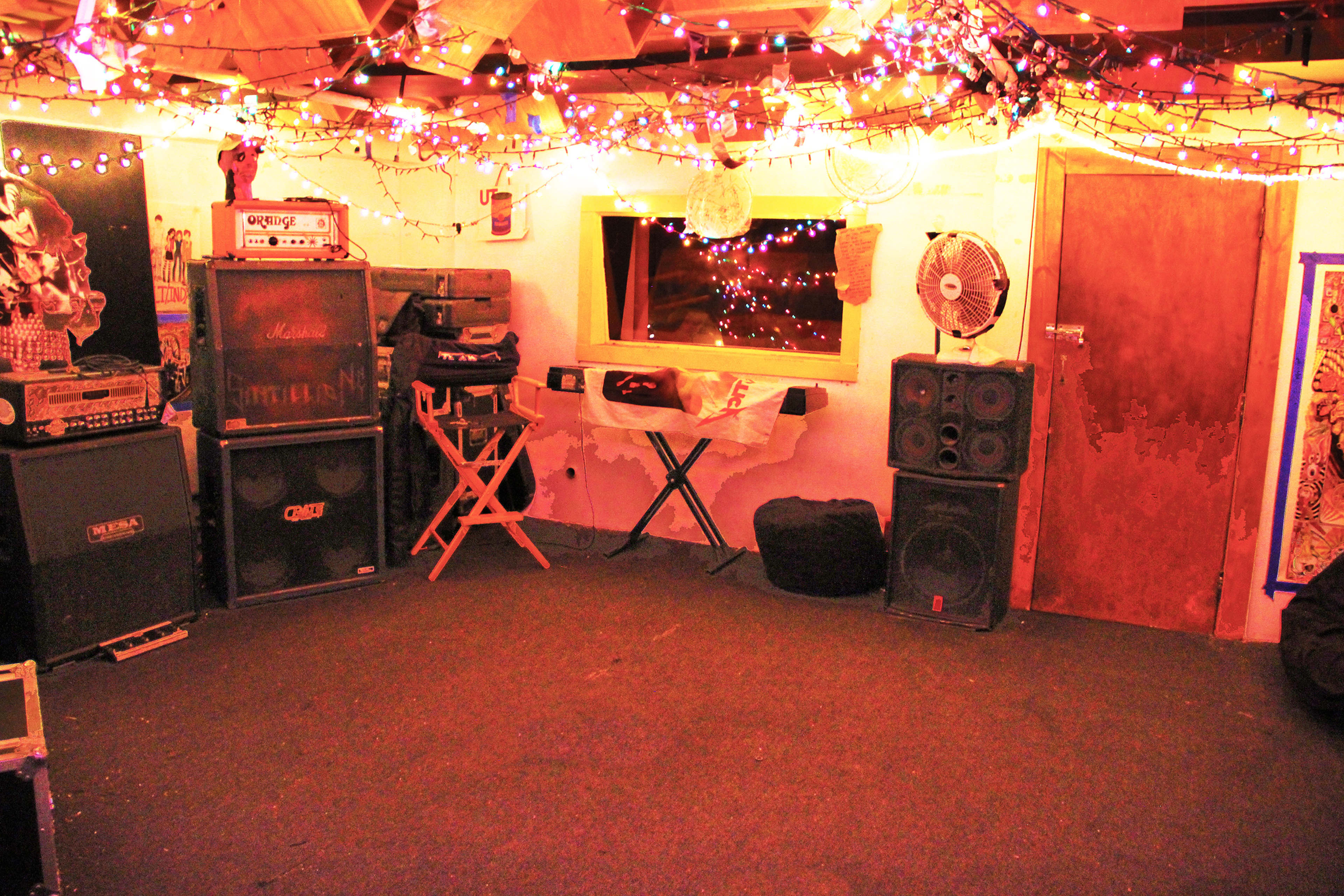 A dimly lit room with colorful string lights overhead, featuring amplifiers and speakers lined against the walls and a window in the back.