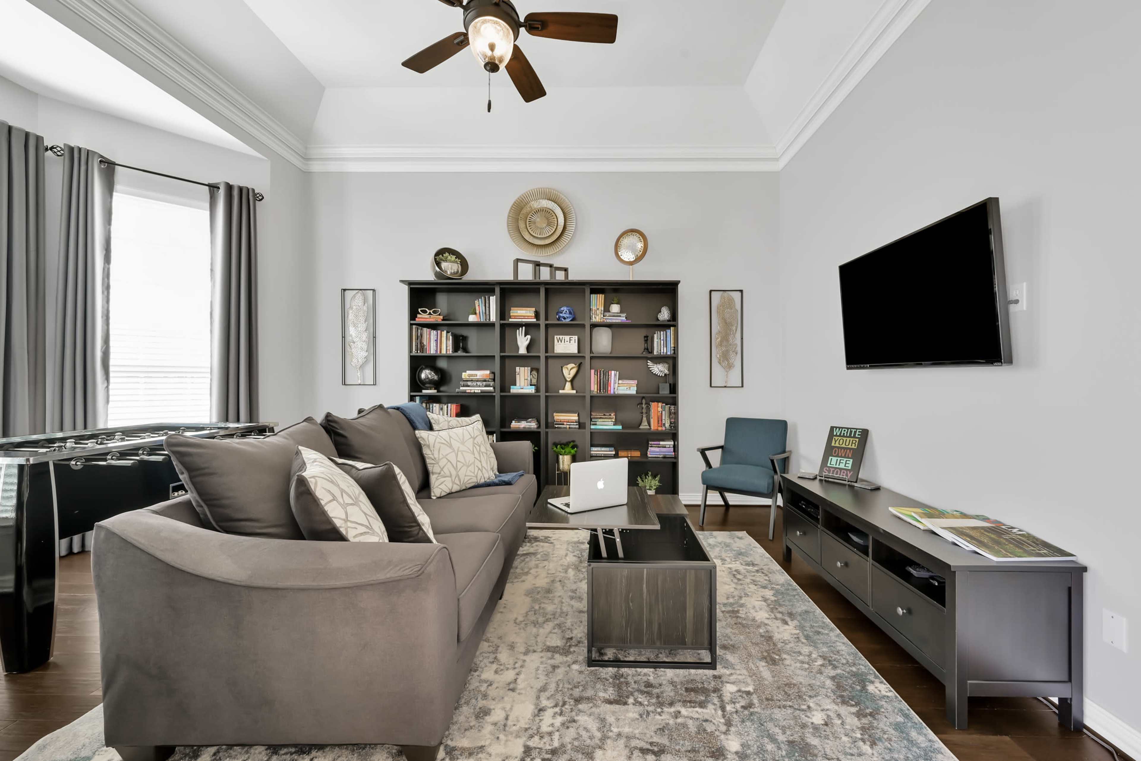 The image shows a modern living room with a grey sofa, a bookshelf filled with books and decorations, and a wall-mounted television.