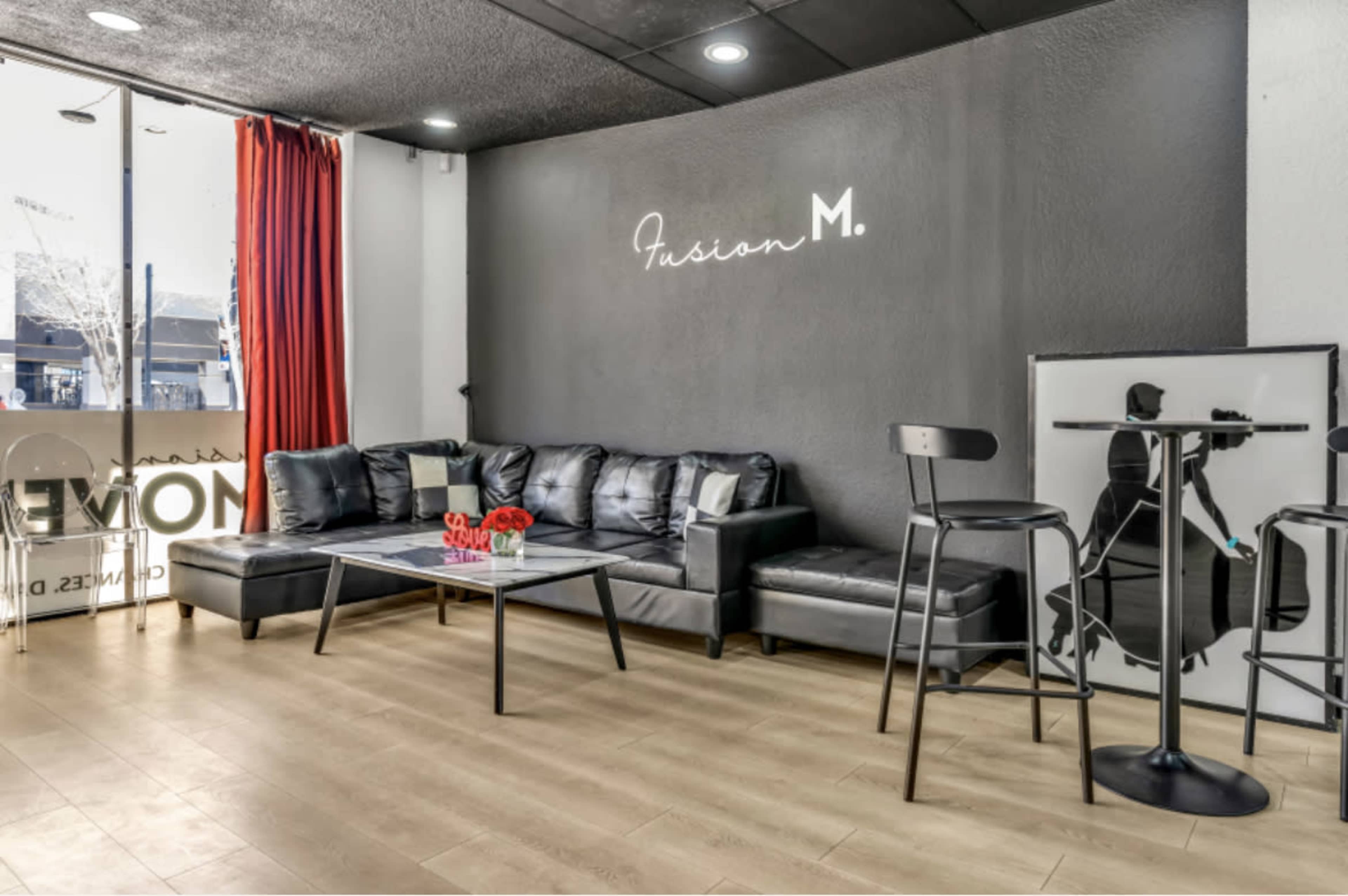 The image features a modern lounge area with a black sectional sofa, a glass coffee table, and bar stools against a dark wall, complemented by red curtains and a decorative logo.