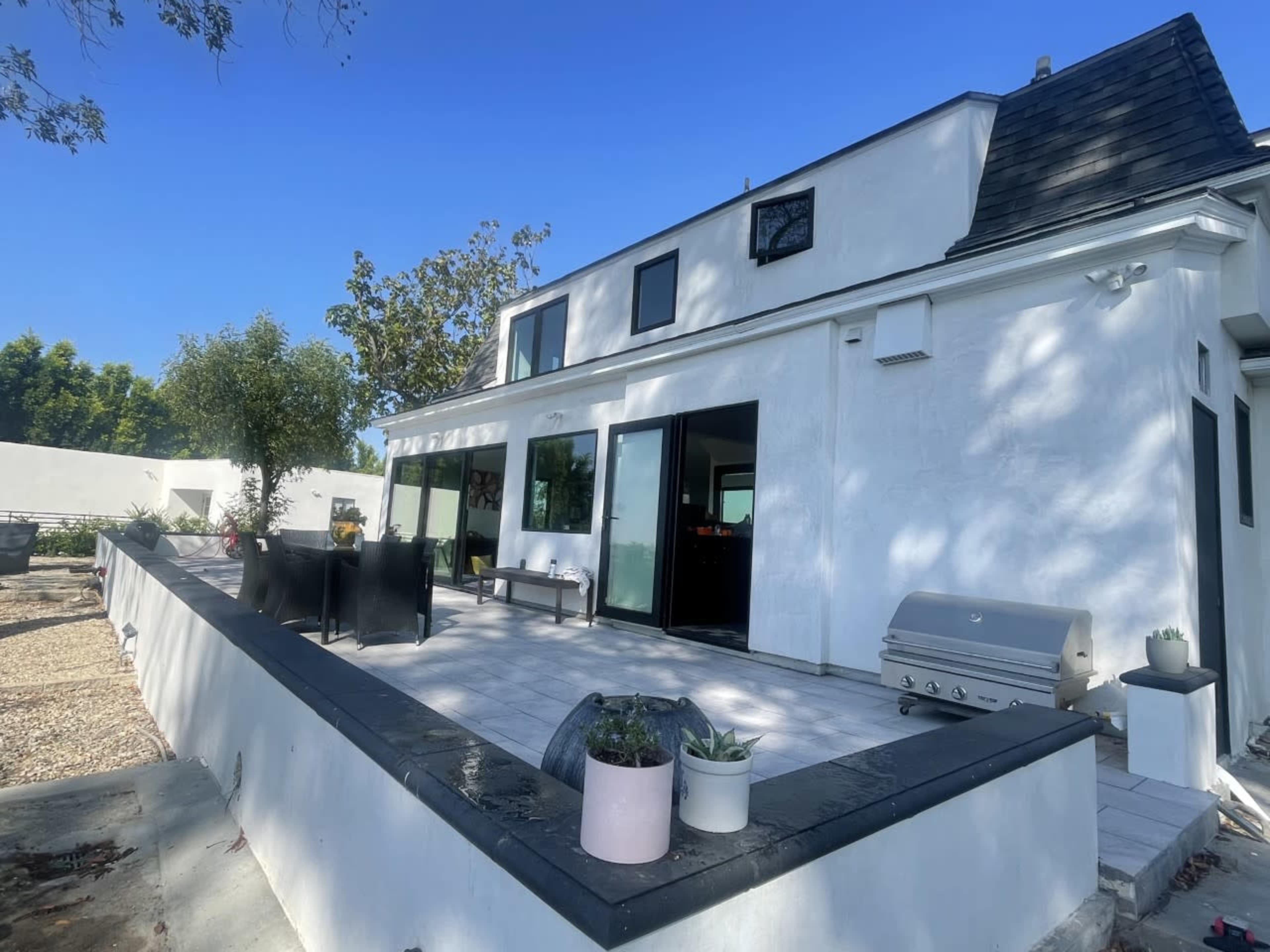 A modern white house with a black roof, large windows, and a patio area featuring outdoor seating and a grill.