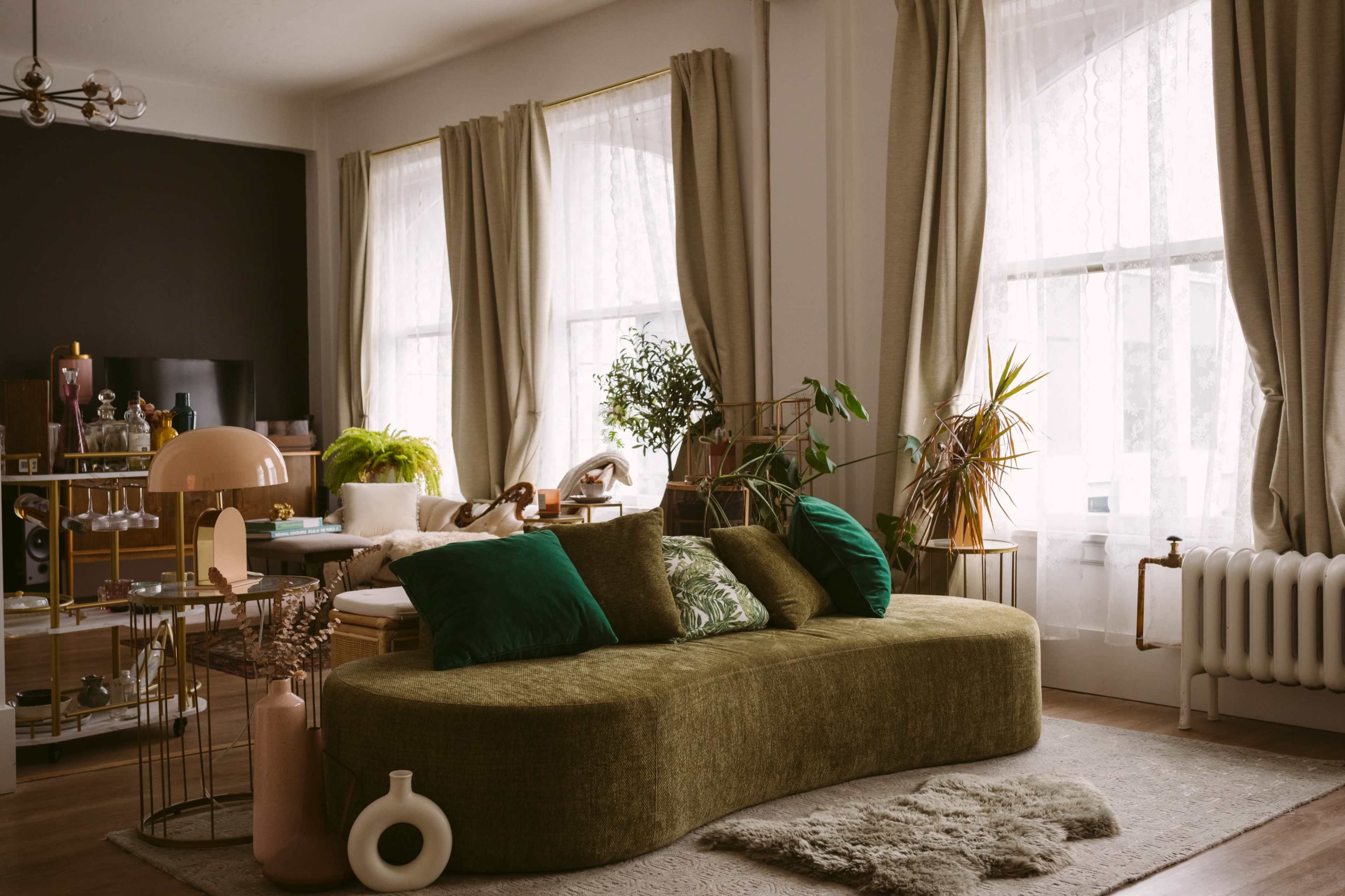 The image shows a modern living room with a curved green sofa, large windows draped with light curtains, and various plants and decorative items arranged around the space.