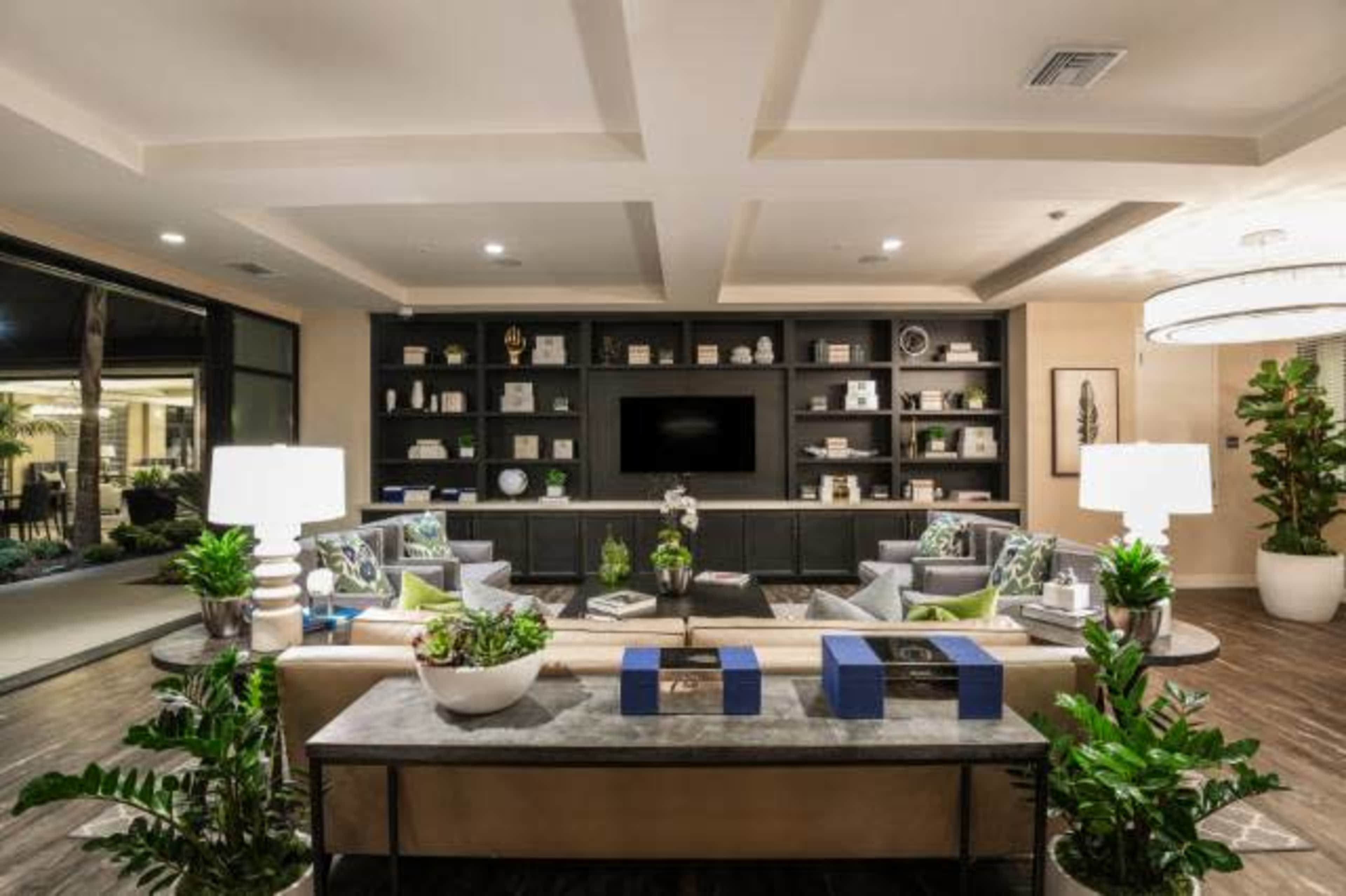 A modern lounge area with a sofa and chairs arranged around a coffee table, flanked by decorative plants and illuminated by table lamps, with a large bookshelf and a television in the background.