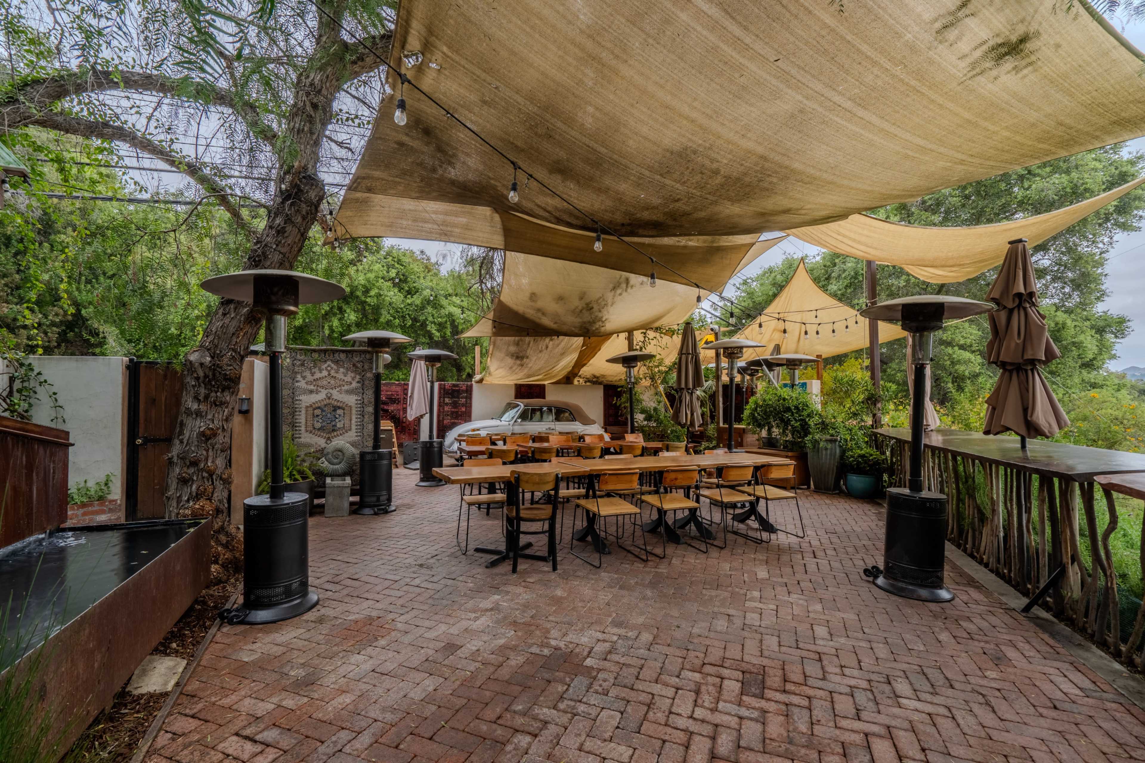 An outdoor dining area features wooden tables and chairs under a canopy of fabric with patio heaters and trees in the background.