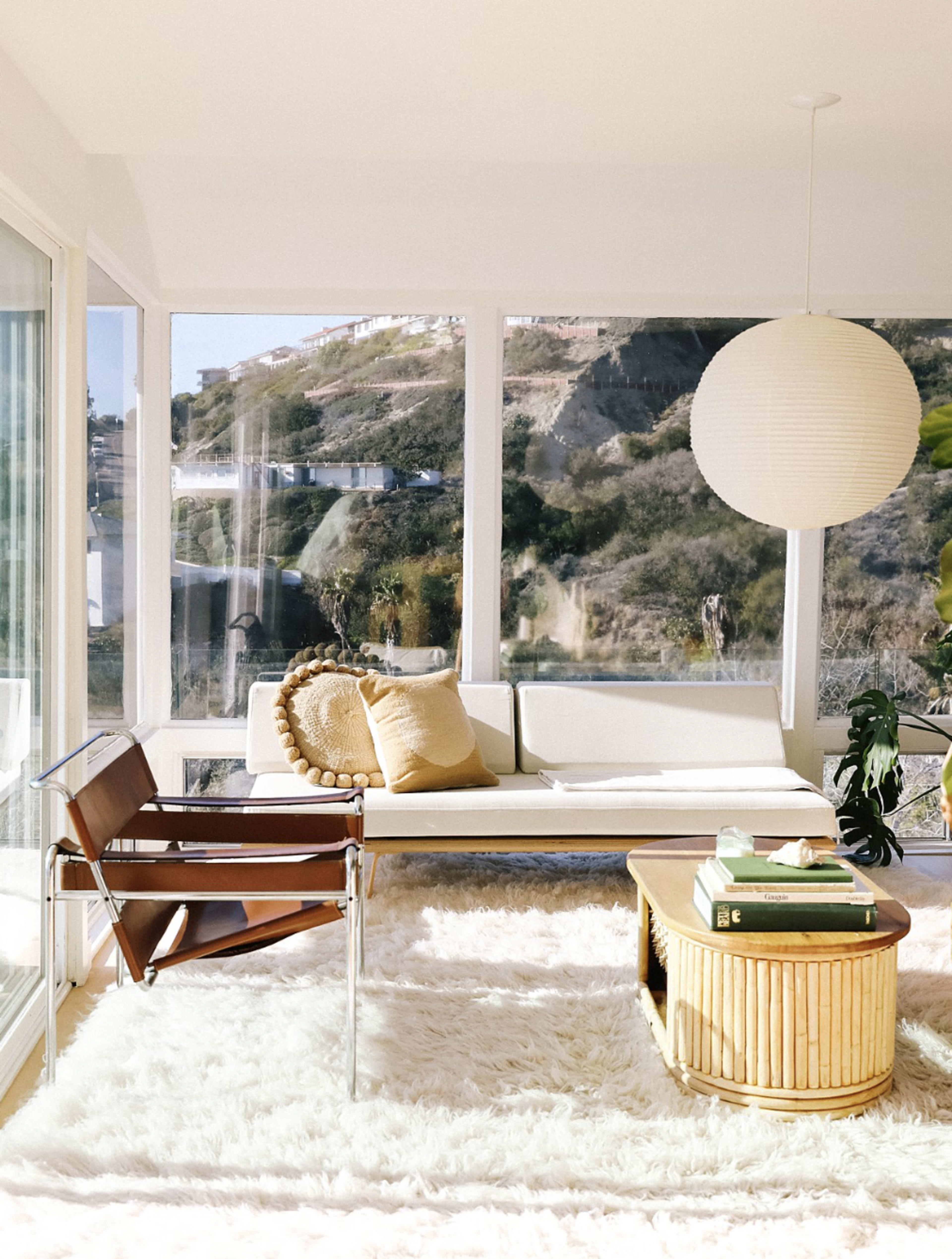 The image shows a bright, modern living room with large windows, featuring a light-colored sofa, a wooden chair, a round pendant lamp, and a circular coffee table on a fluffy rug.