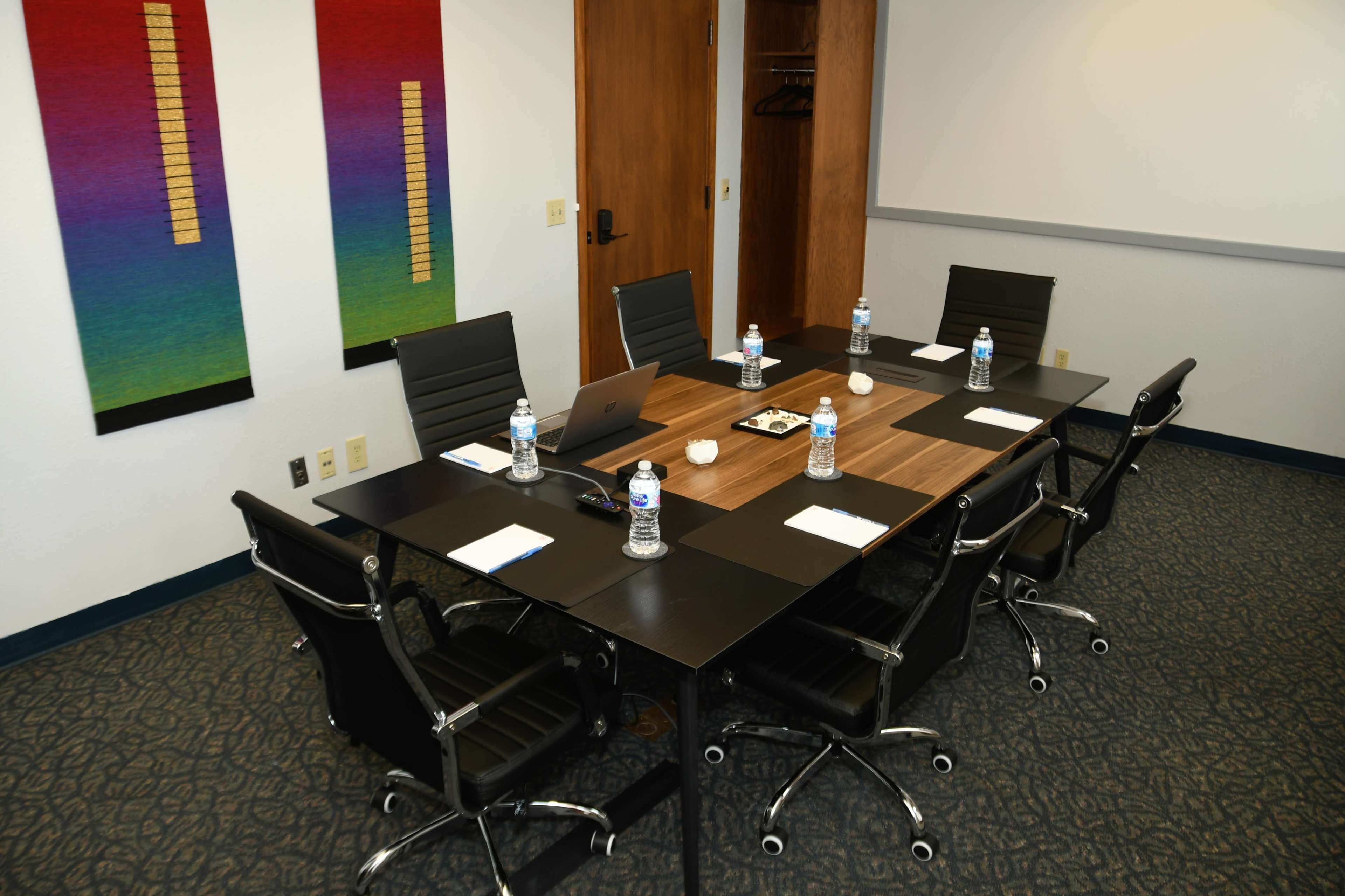 A modern conference room with a rectangular table, six black chairs, and water bottles placed at each seat, flanked by colorful wall art.