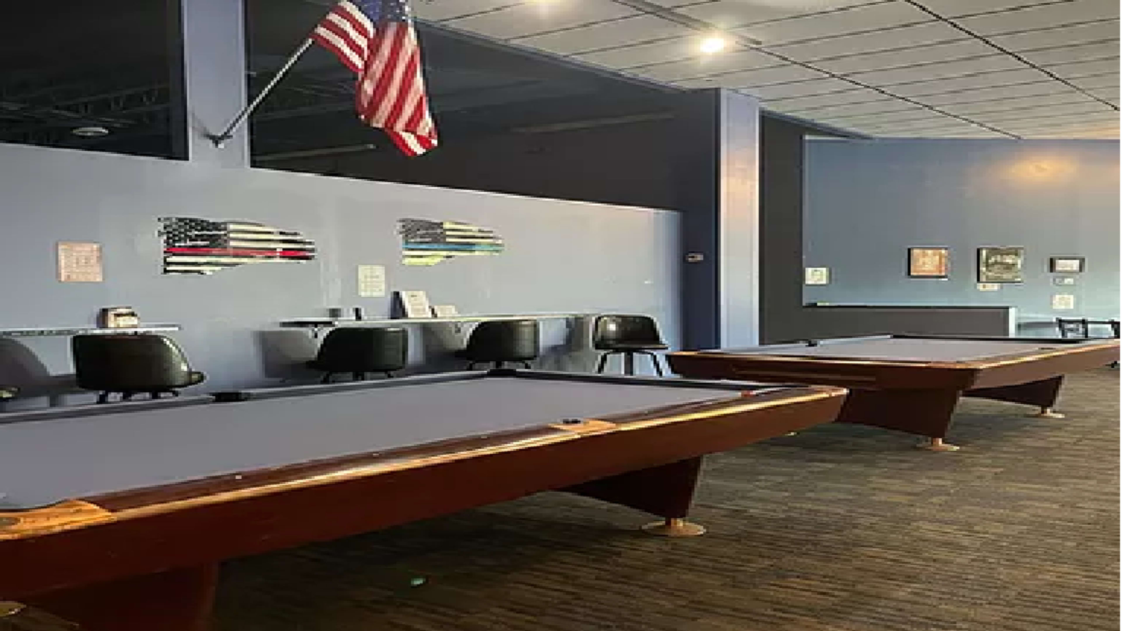 A room with two pool tables, an American flag hanging from the ceiling, and a few chairs lined against the wall.