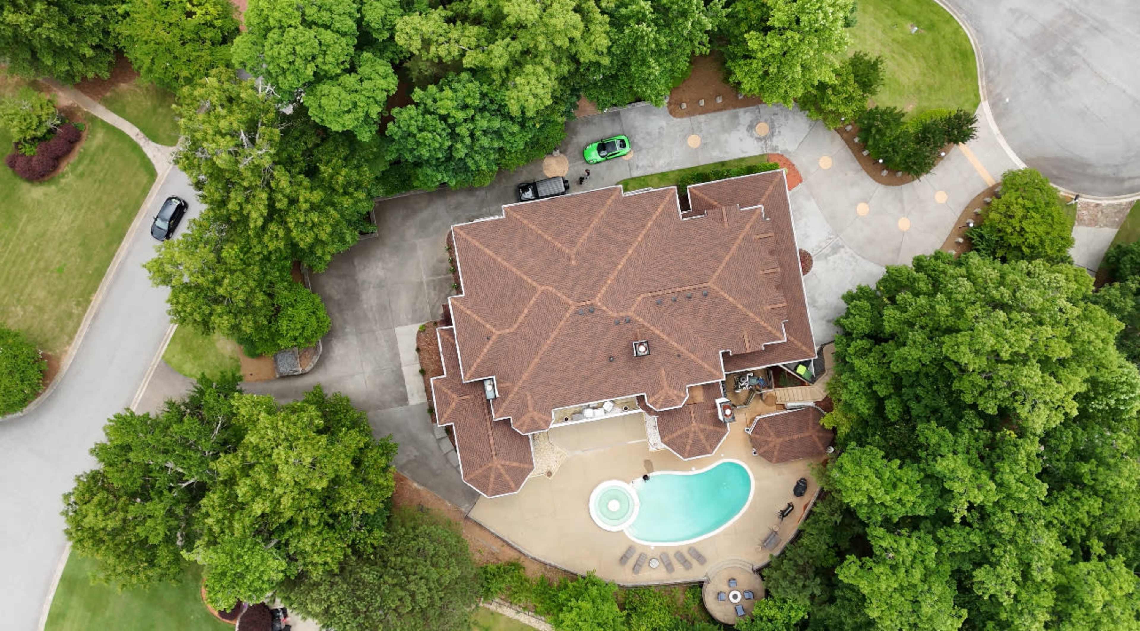 A large house with a swimming pool surrounded by trees and a paved driveway, captured from an aerial perspective.