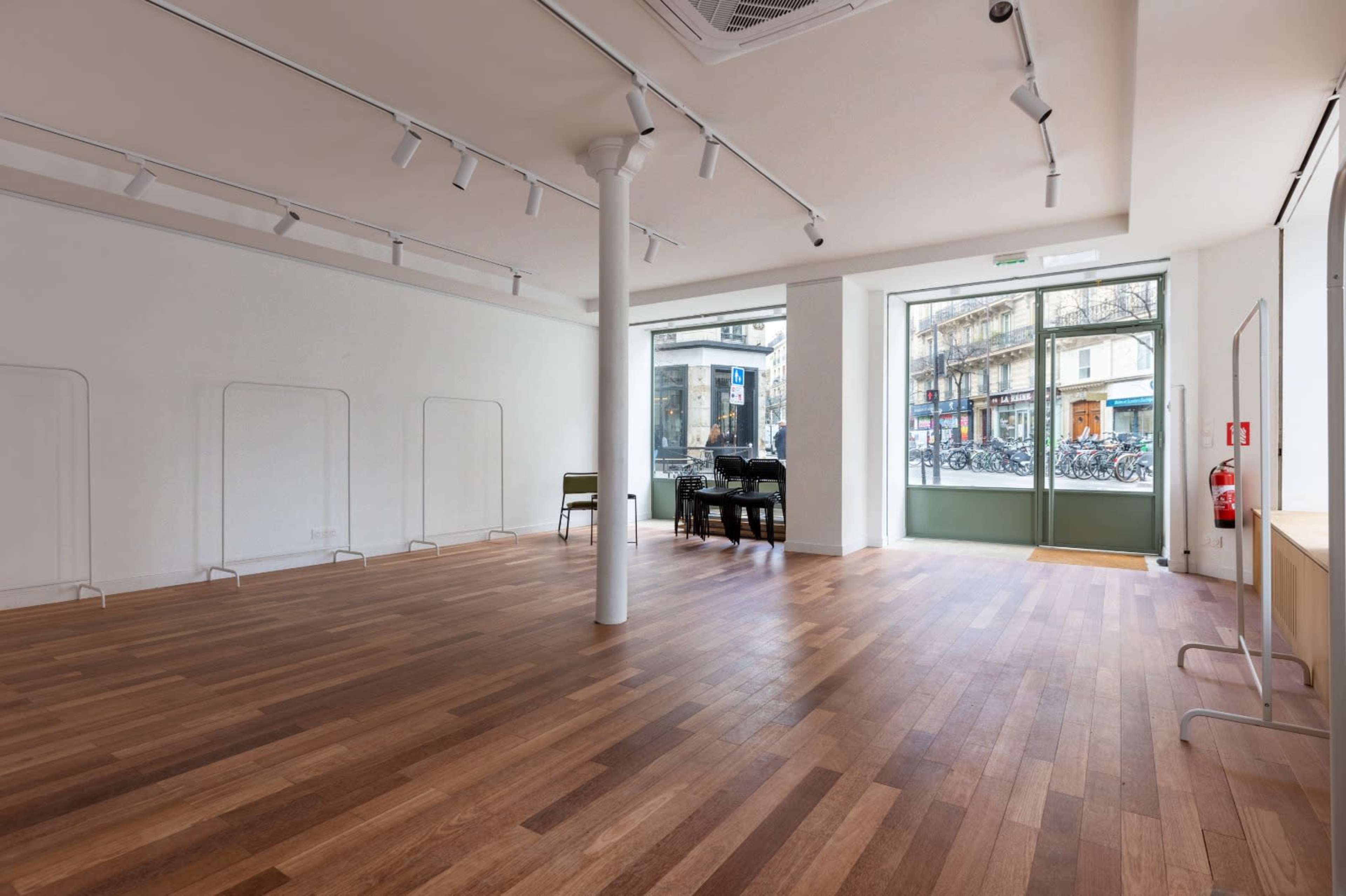 The image shows a spacious, empty interior room with wooden flooring and large windows overlooking a street with bicycles parked outside.