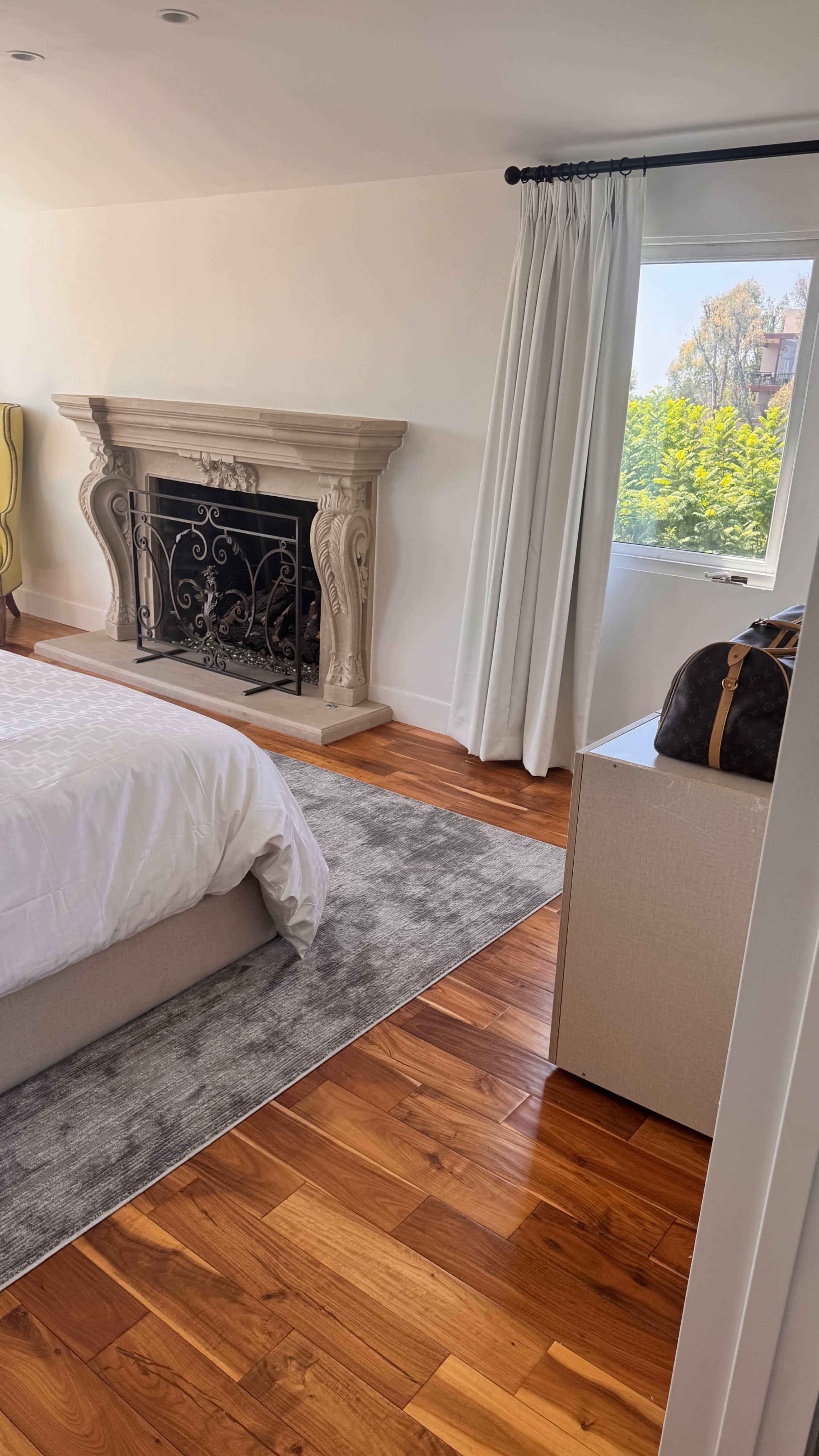 The image shows a modern bedroom featuring a bed, a decorative fireplace with ornate details, and a window with light curtains.