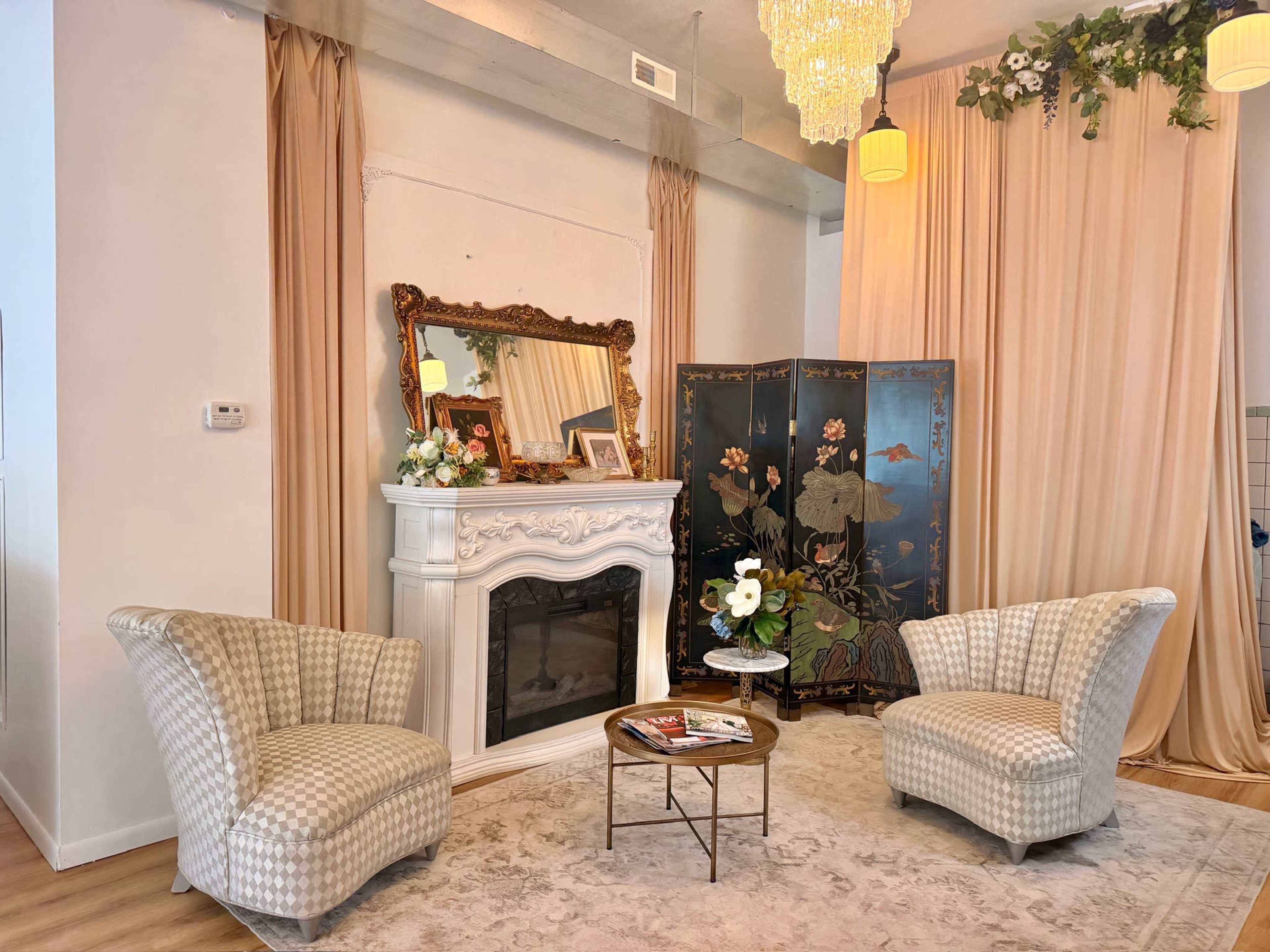 The scene features two patterned armchairs facing a decorative fireplace, with a mirror above it, flanked by a folding screen and draped curtains in a spacious room.