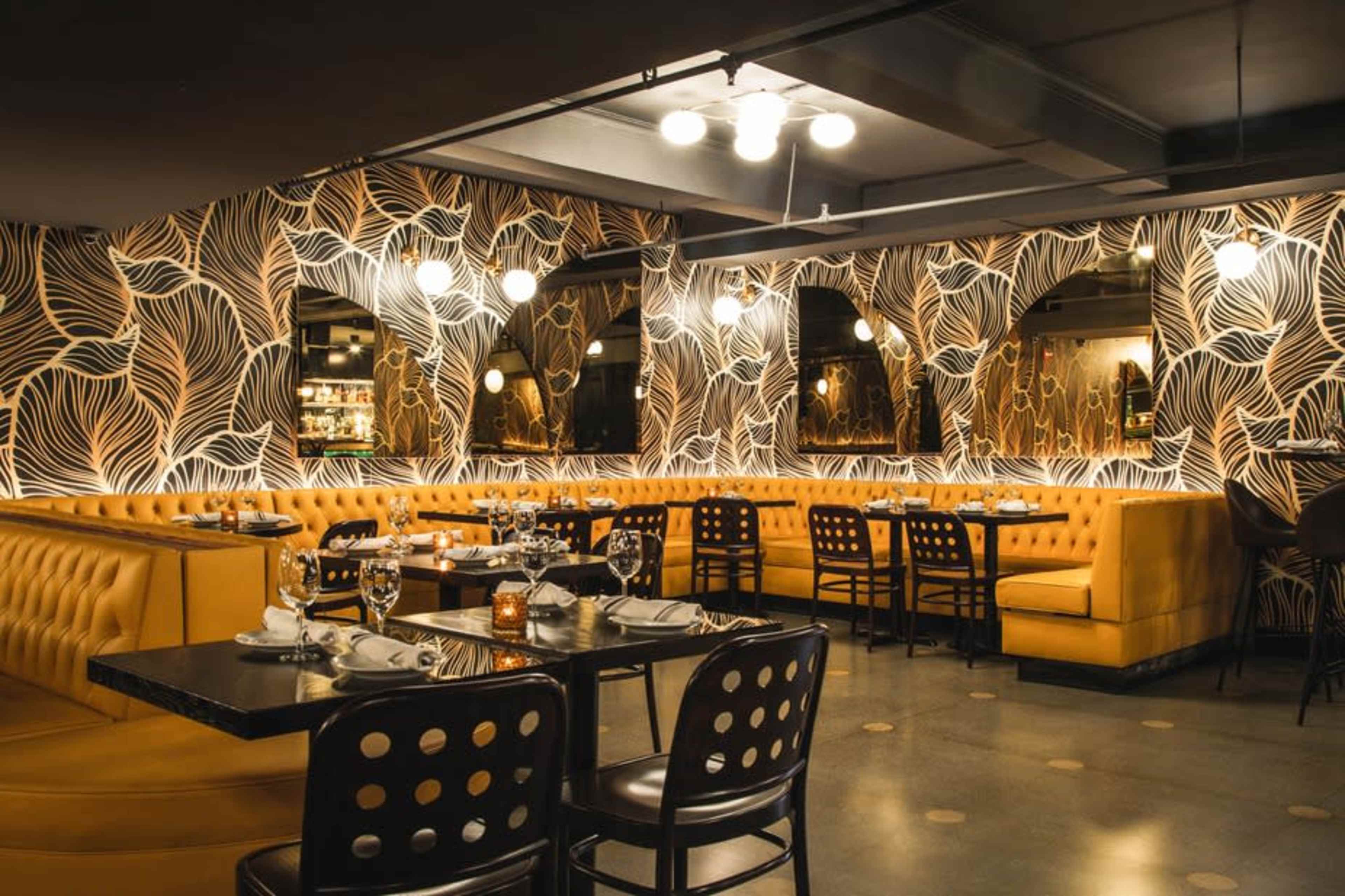 The image shows the interior of a restaurant featuring a lively wallpaper design, yellow upholstered seating, and dark wooden tables with black chairs.