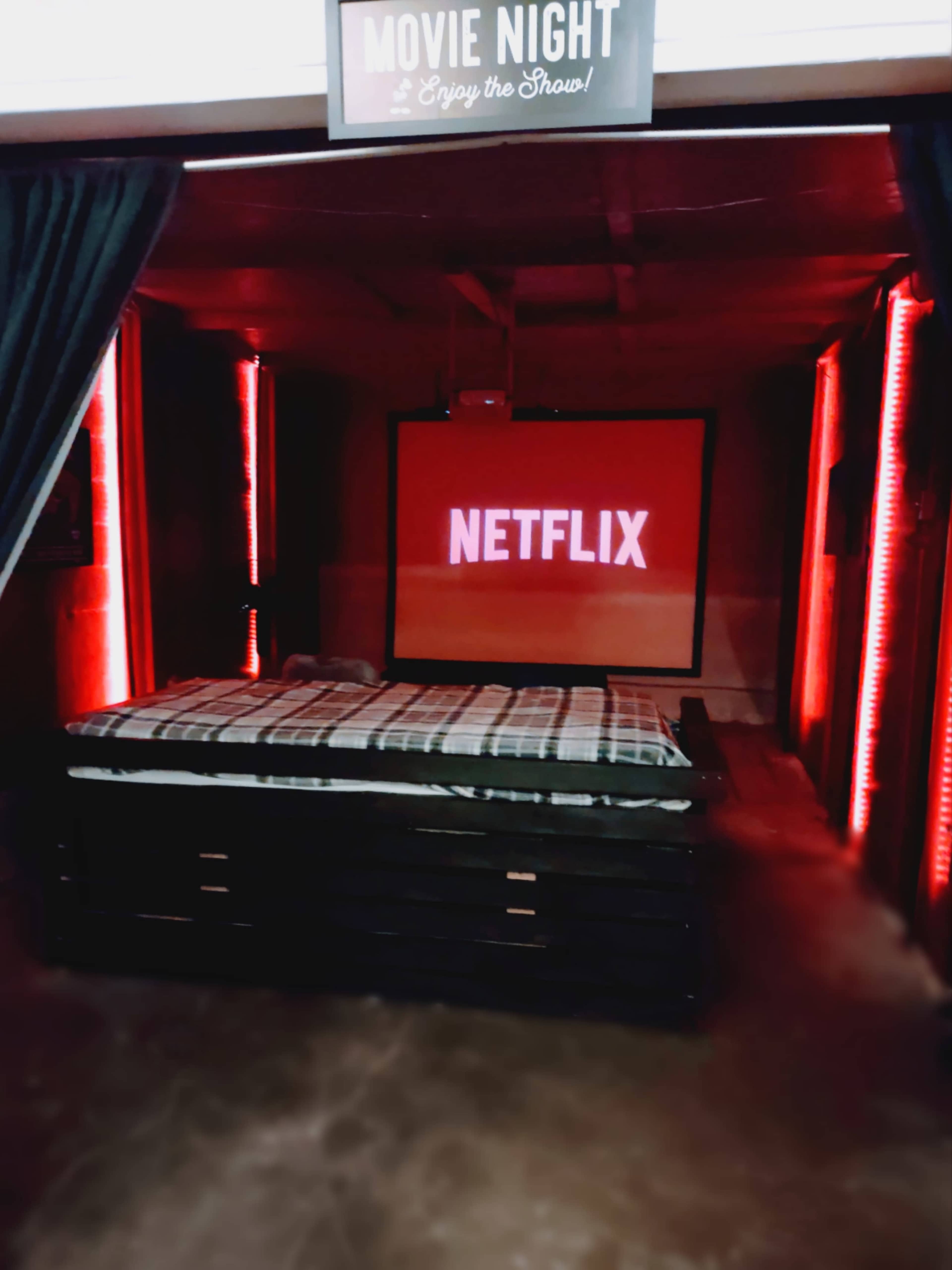 A cozy movie night setup featuring a large screen displaying the Netflix logo, a bed with a checkered blanket, and red LED lights illuminating the space.