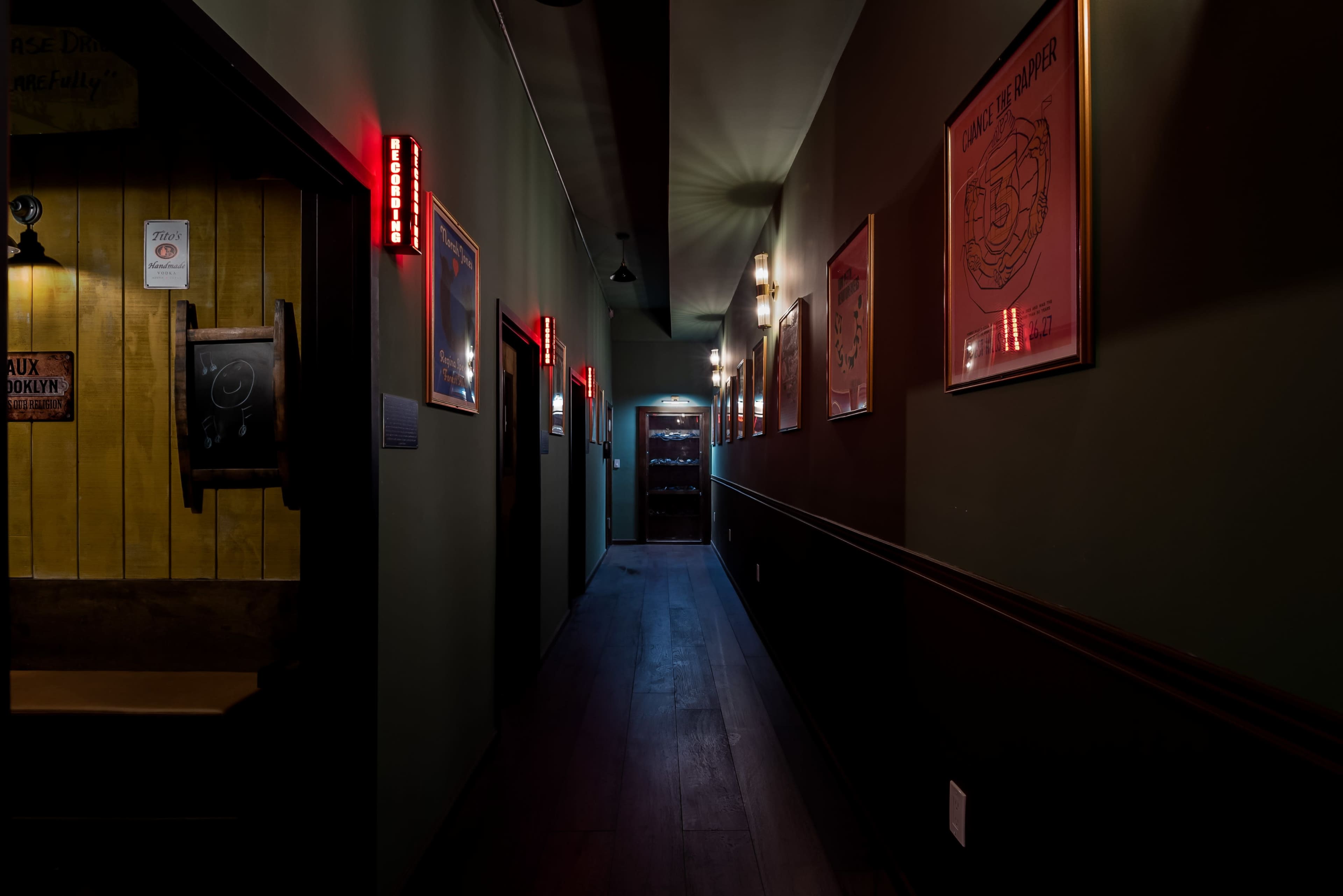 A narrow hallway with dark green walls is illuminated by red and white lights, featuring framed artwork on the sides and a door at the end.