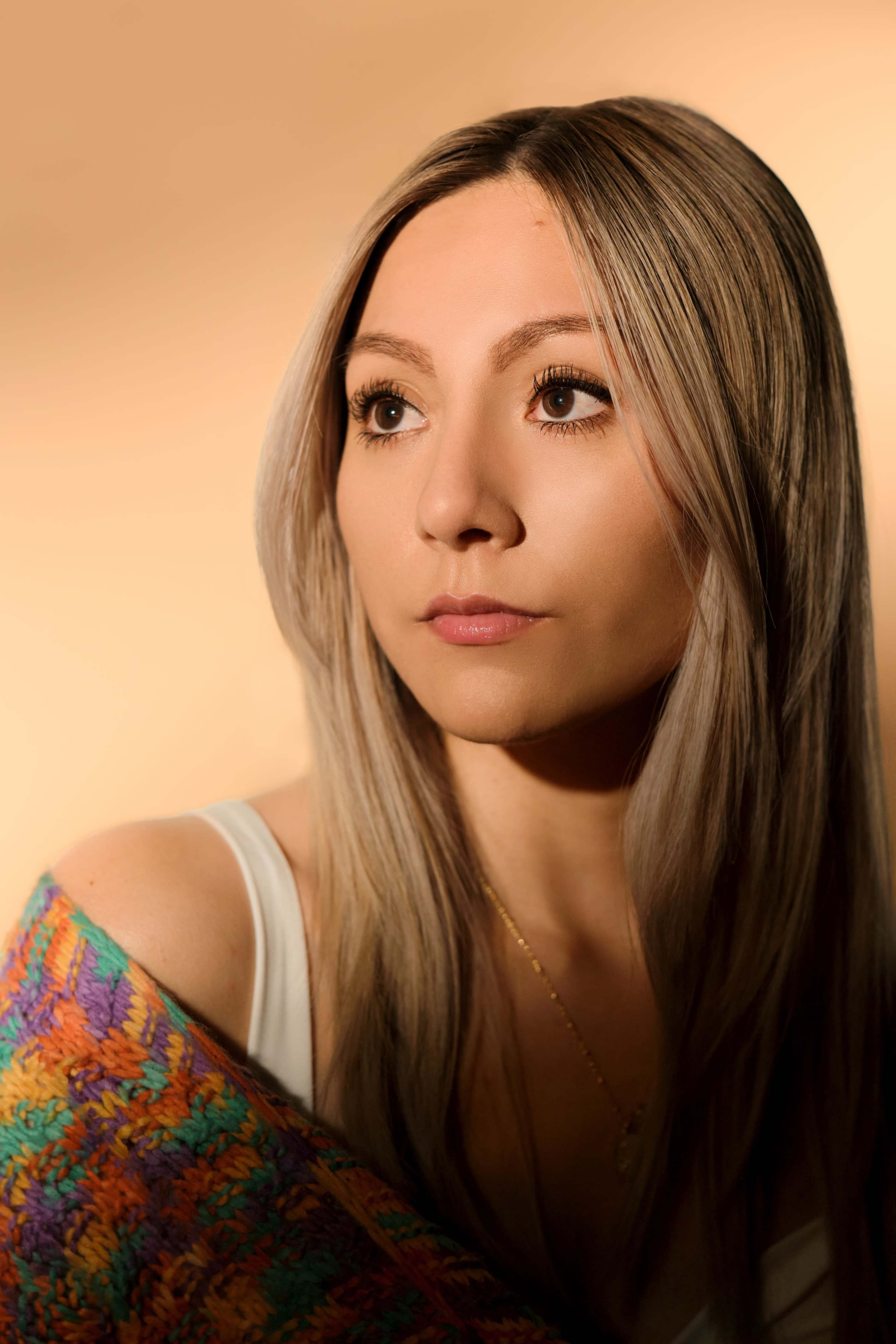 A woman with long, light hair gazes softly to the side while draped in a colorful shawl against a warm background.
