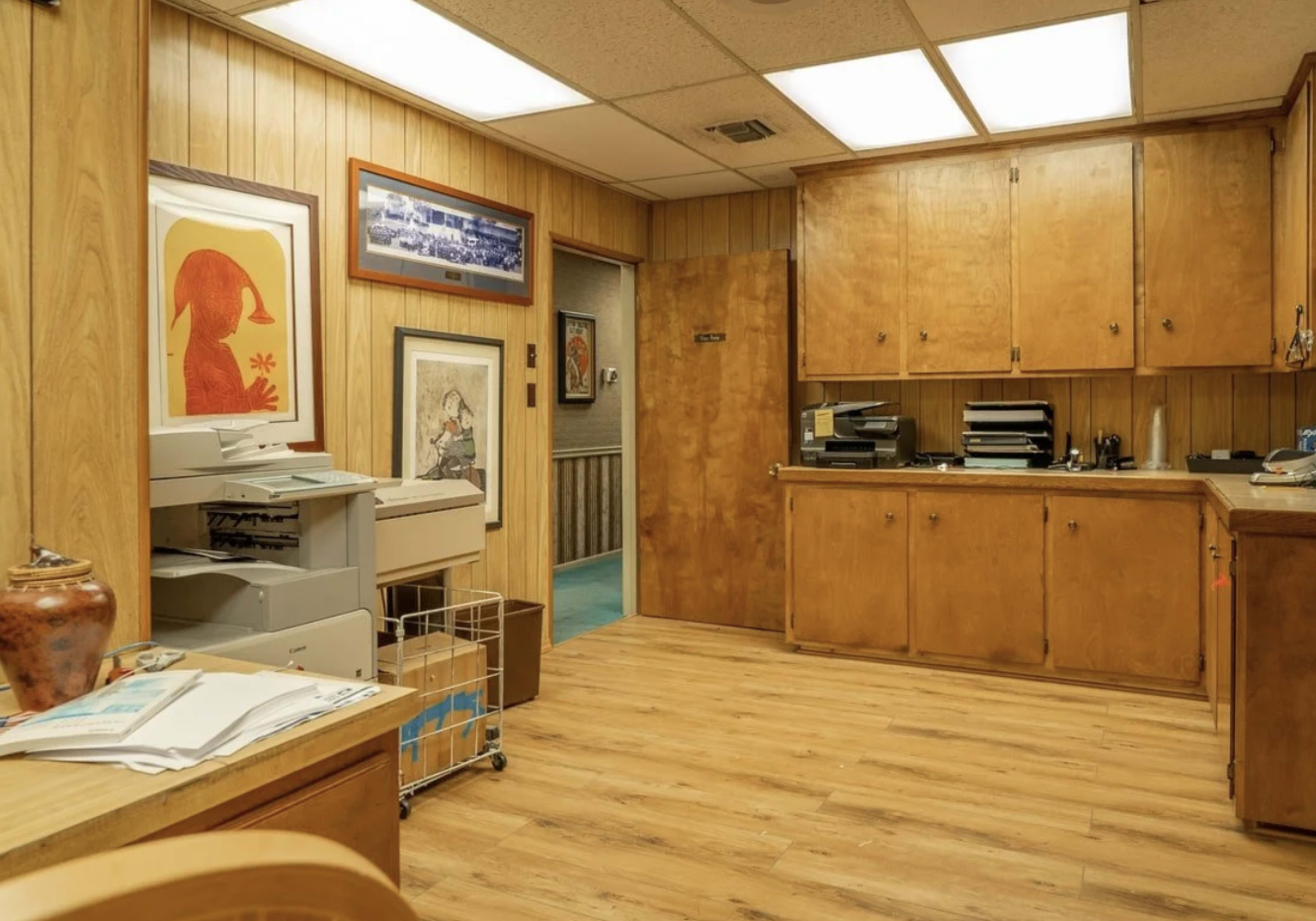 The image shows an office space with wooden paneling, a desk, a printer, and various framed artworks on the walls.
