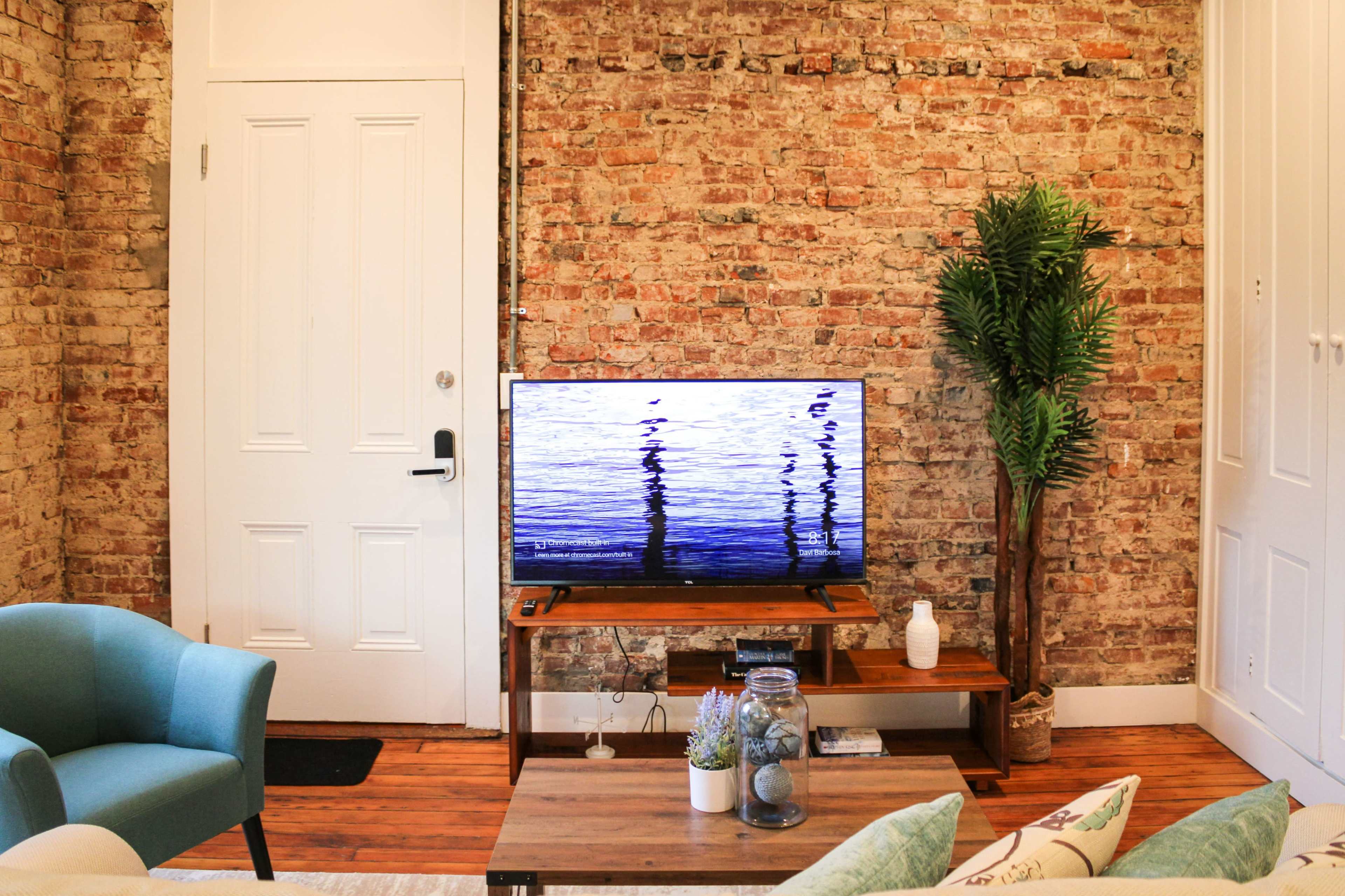 A television displaying a water reflection is positioned on a wooden stand against a brick wall in a room featuring a green armchair and a potted plant.