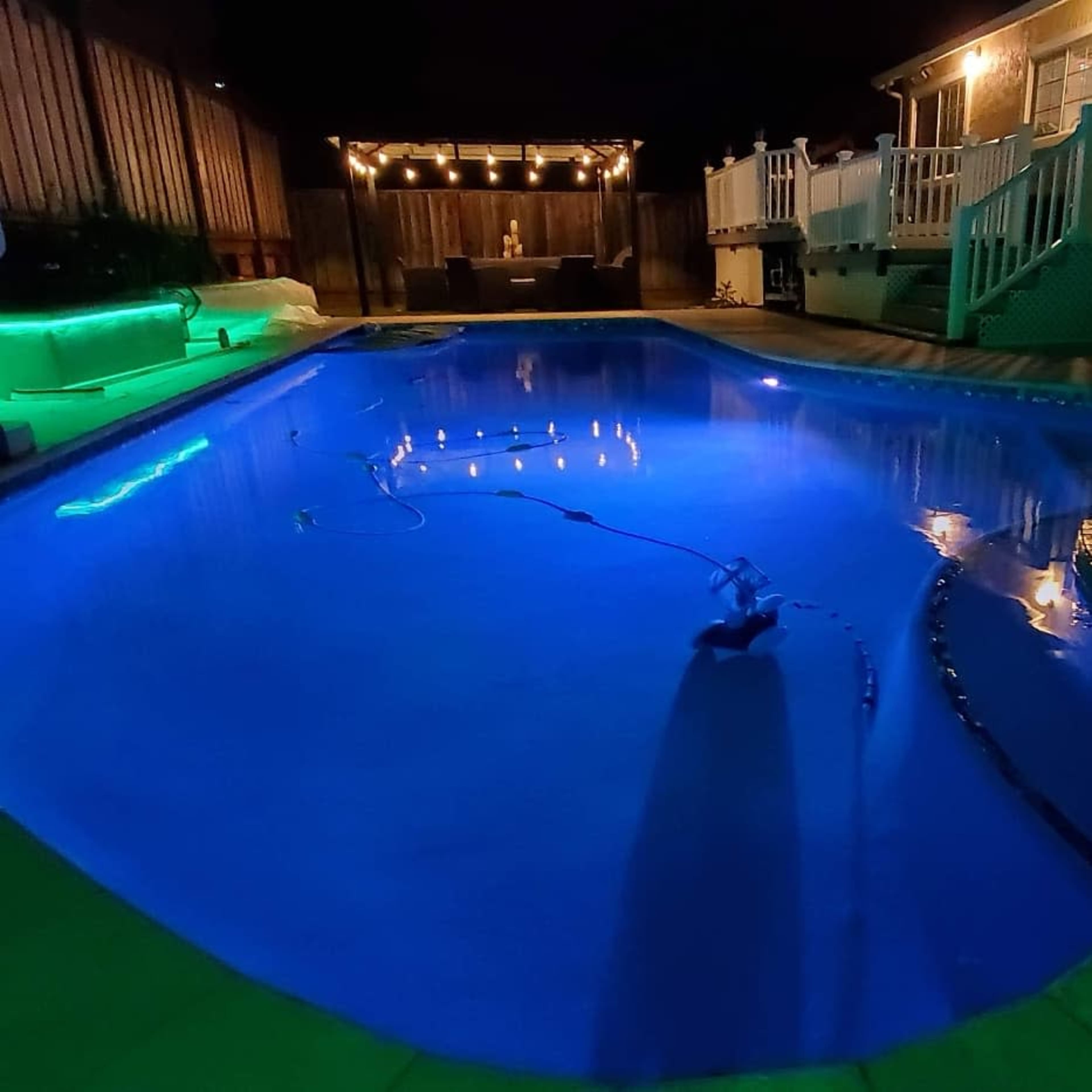 A well-lit swimming pool at night with a patio area featuring string lights in the background.