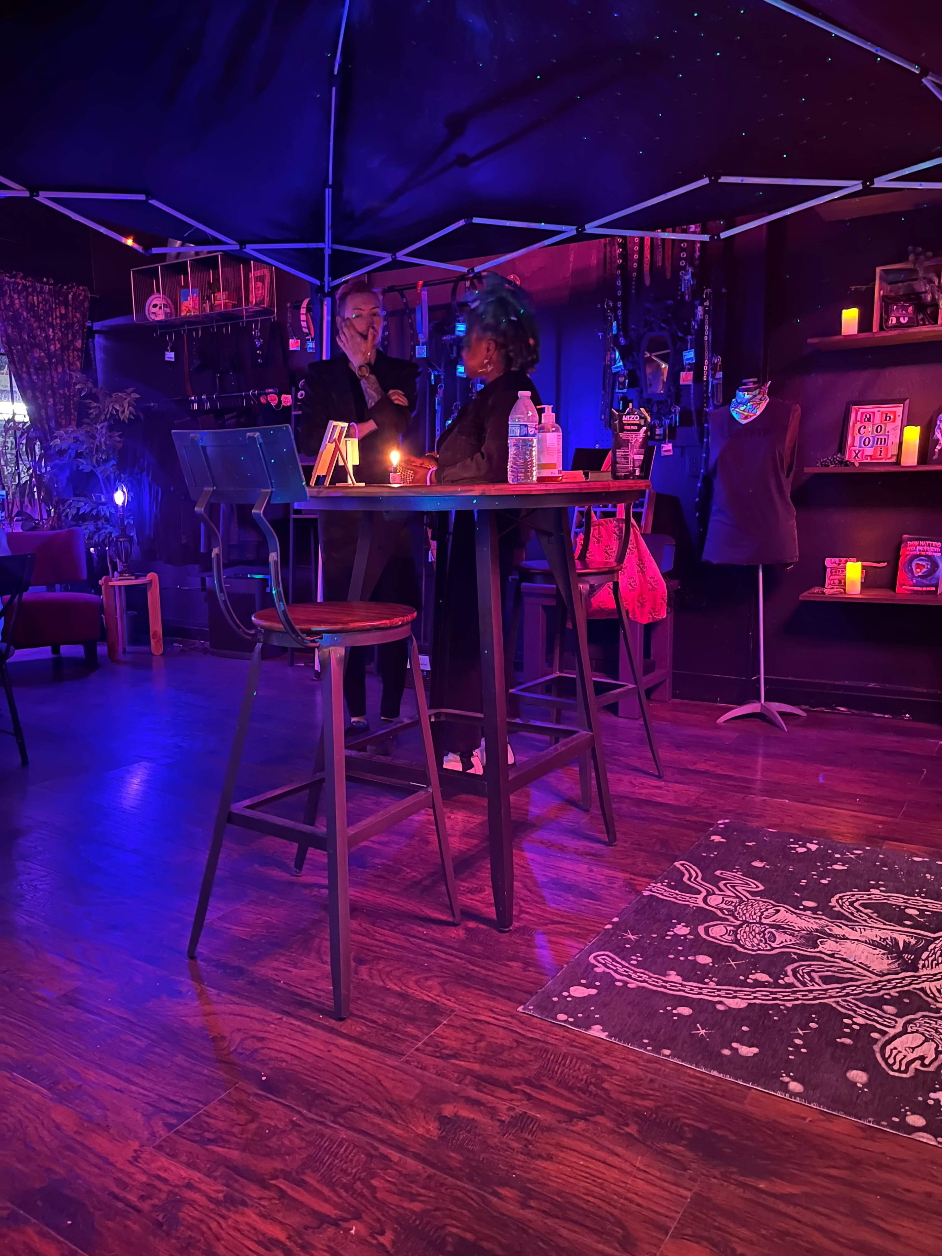 A dimly lit lounge features a high bar table with two people standing nearby, surrounded by colorful decorations and shelves filled with items.
