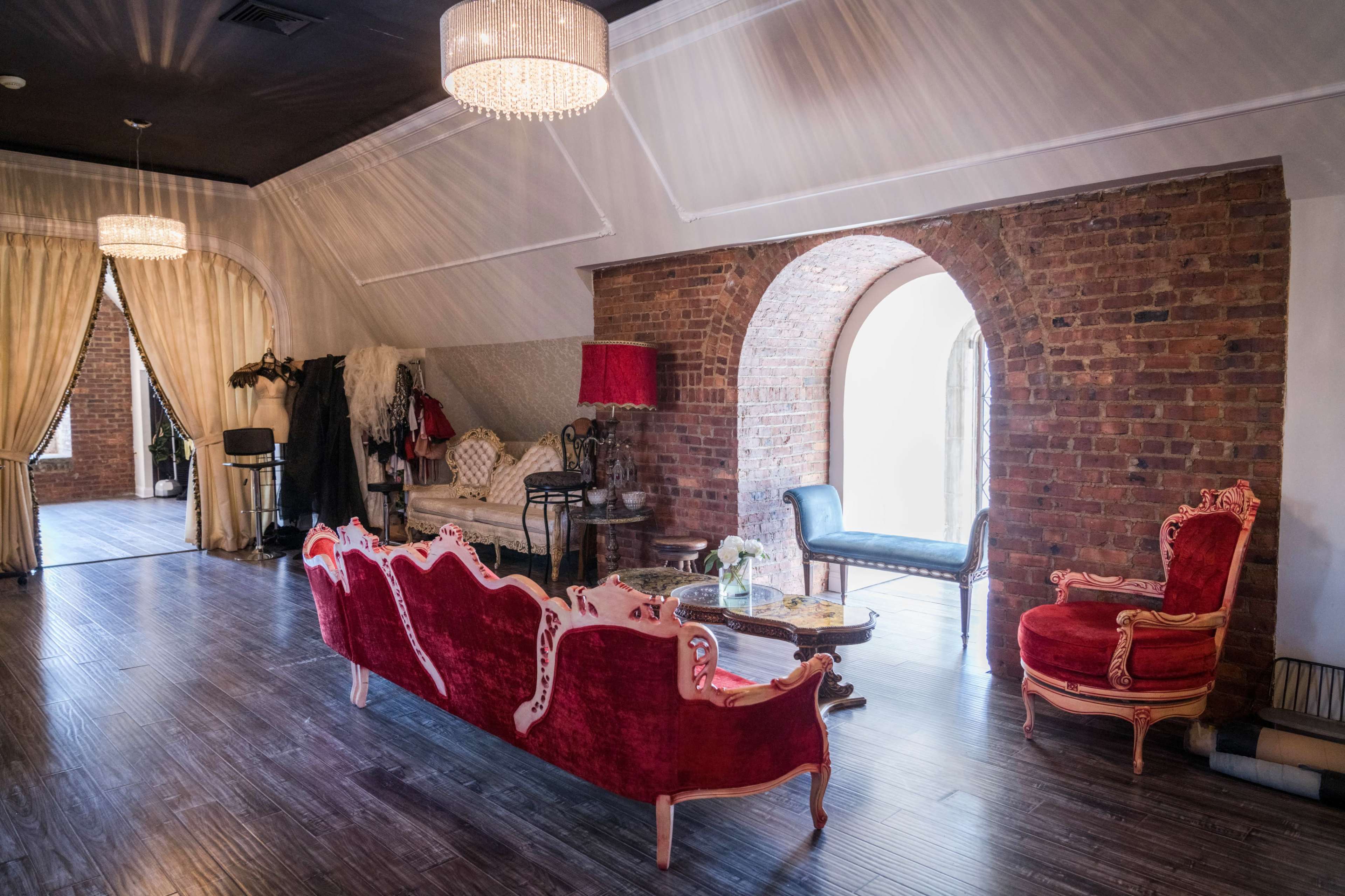A spacious room with brick walls, elegant vintage furniture, and large arched windows, illuminated by chandeliers.