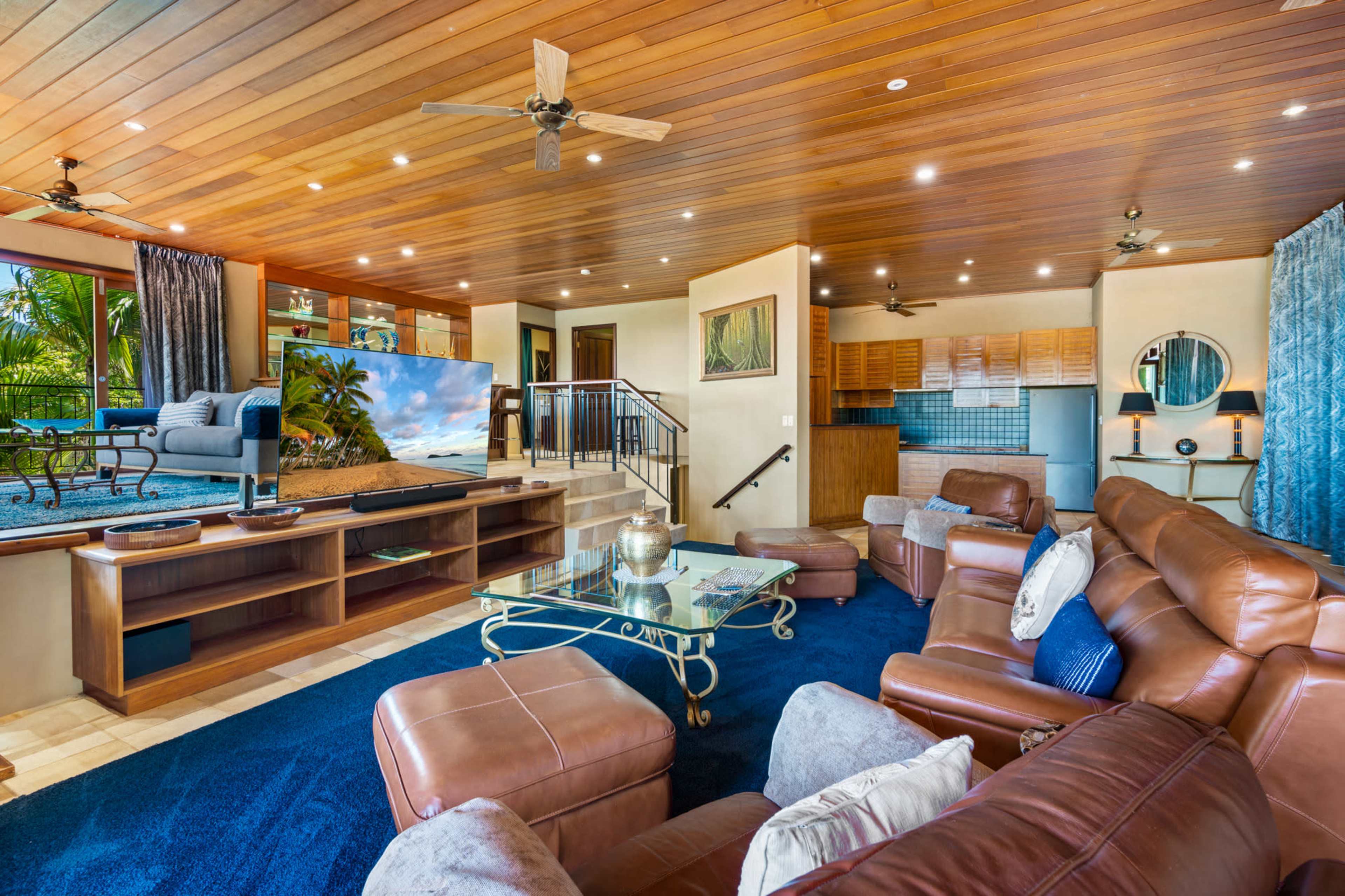 A spacious living room features leather seating, a large TV, and a view of tropical greenery through expansive windows.