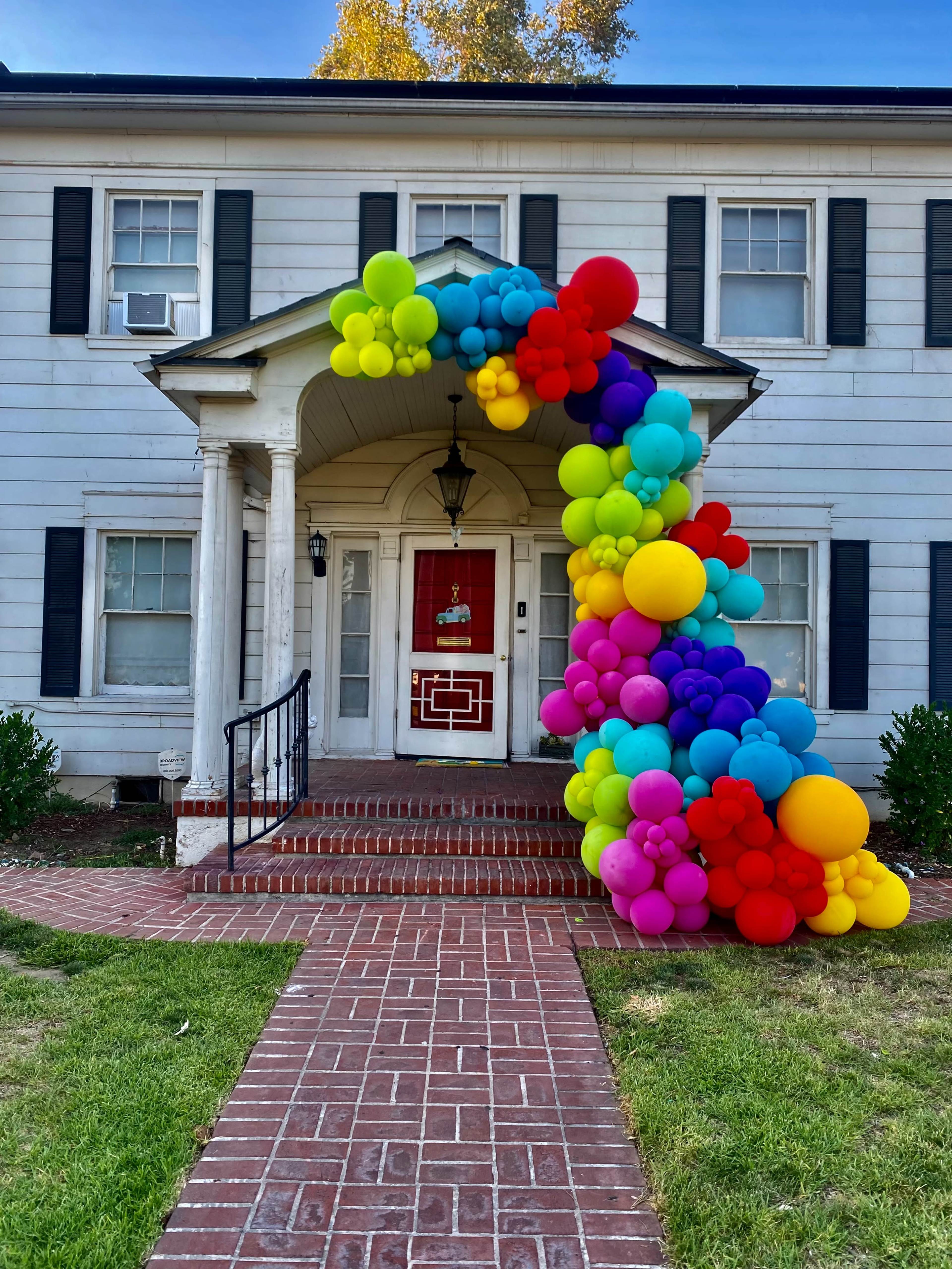 A colorful arrangement of balloons in various sizes adorns the entrance of a white house with a red door.