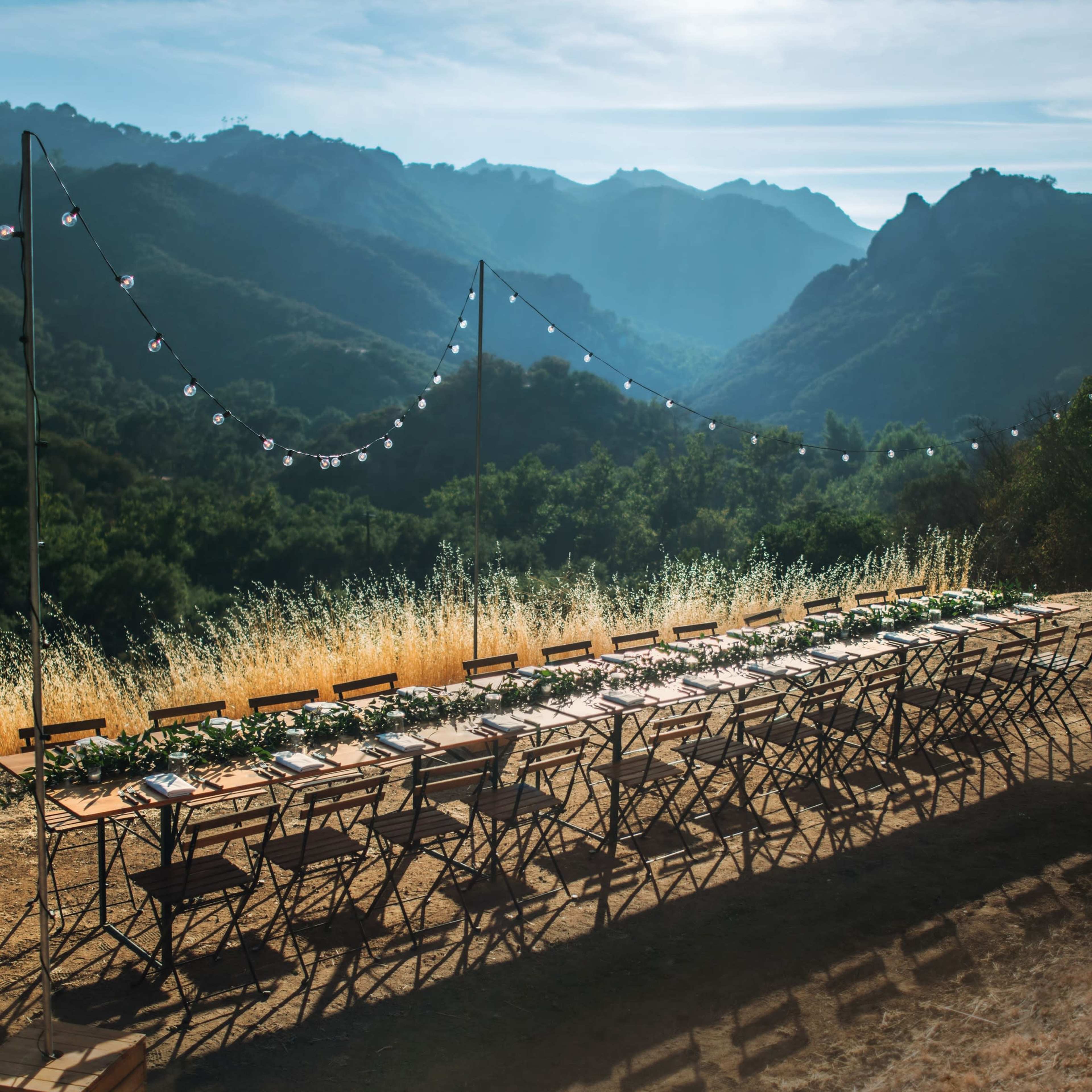 A long wooden table is set for a meal outdoors, surrounded by folding chairs, with mountains and a clear sky in the background.