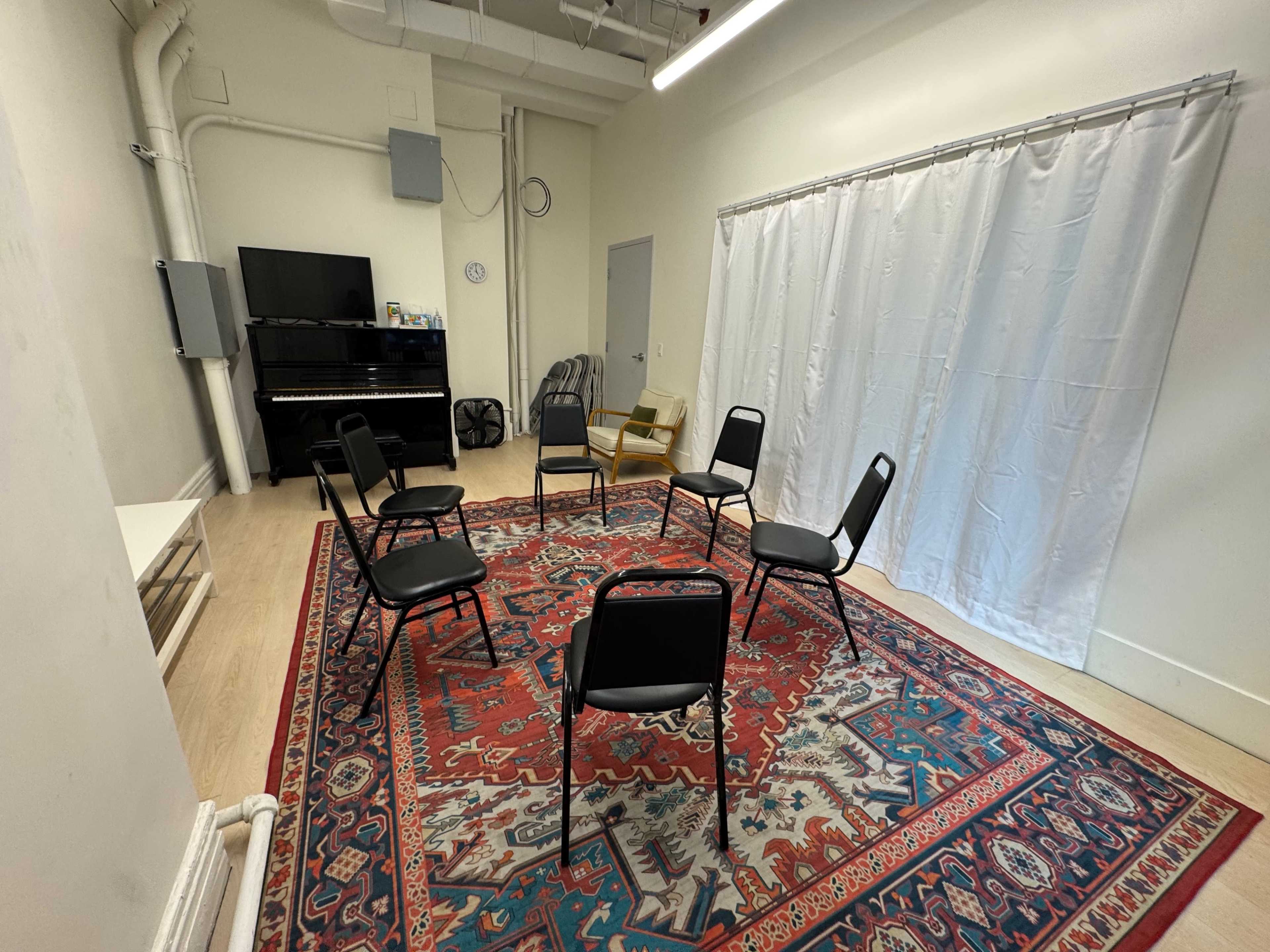 The image shows a small room with a large patterned rug, six chairs arranged in a circle, a white curtain, and a piano against one wall.