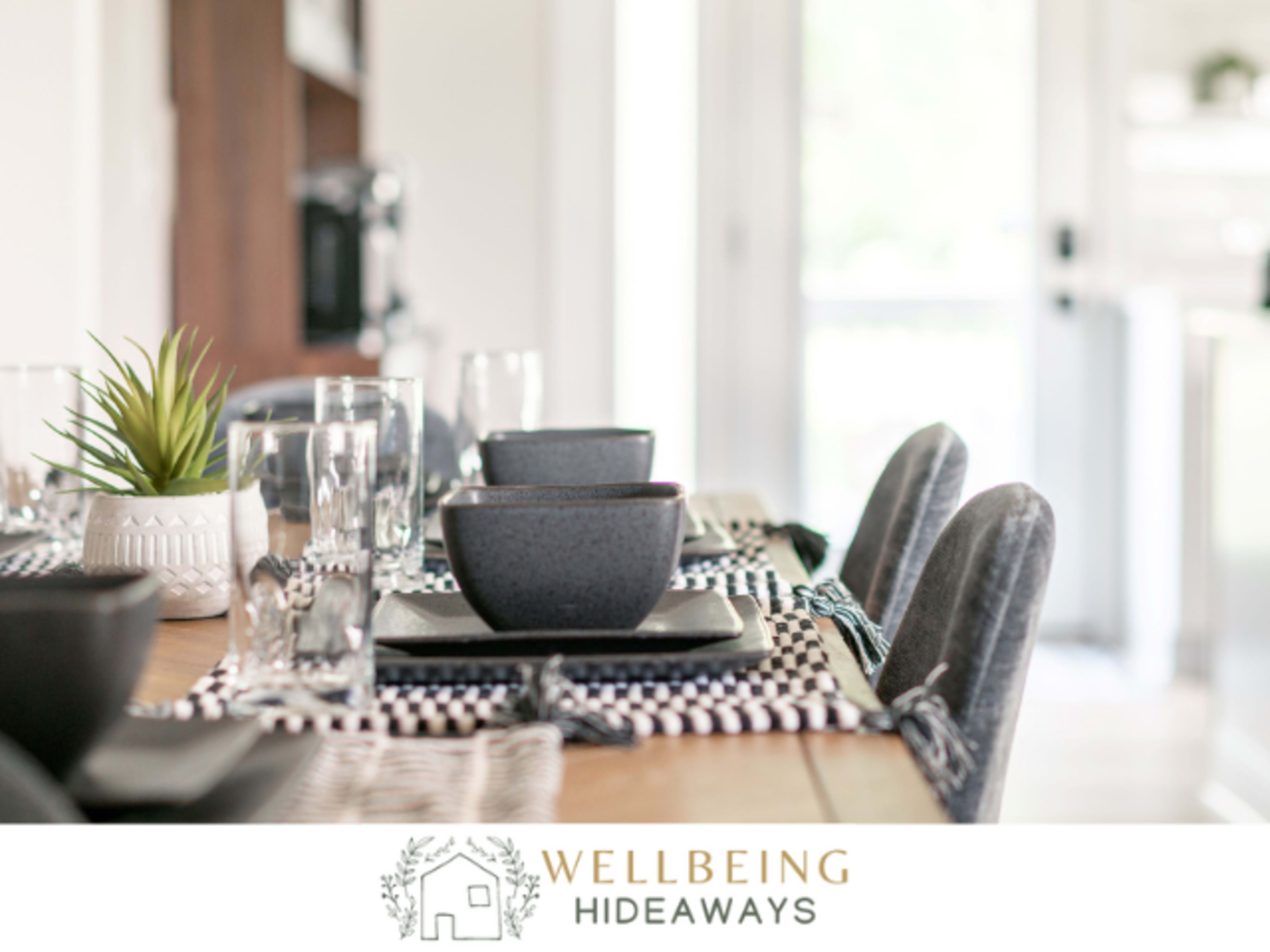 A dining table is set with black bowls, clear glasses, and placemats, complemented by a small green plant in a white pot.