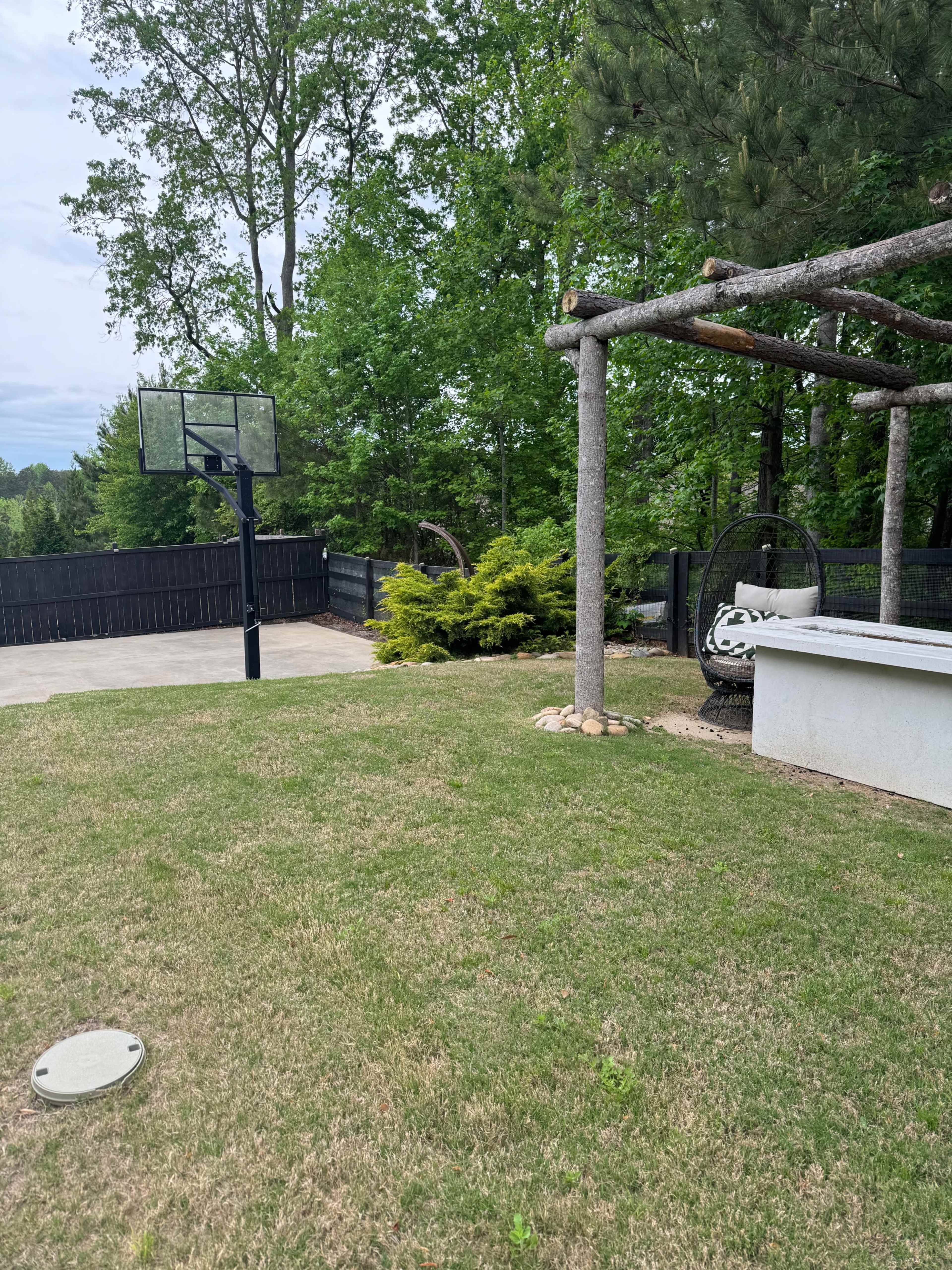 The image shows a backyard with a basketball hoop, a gravel area with a bench, and lush greenery surrounding the space.