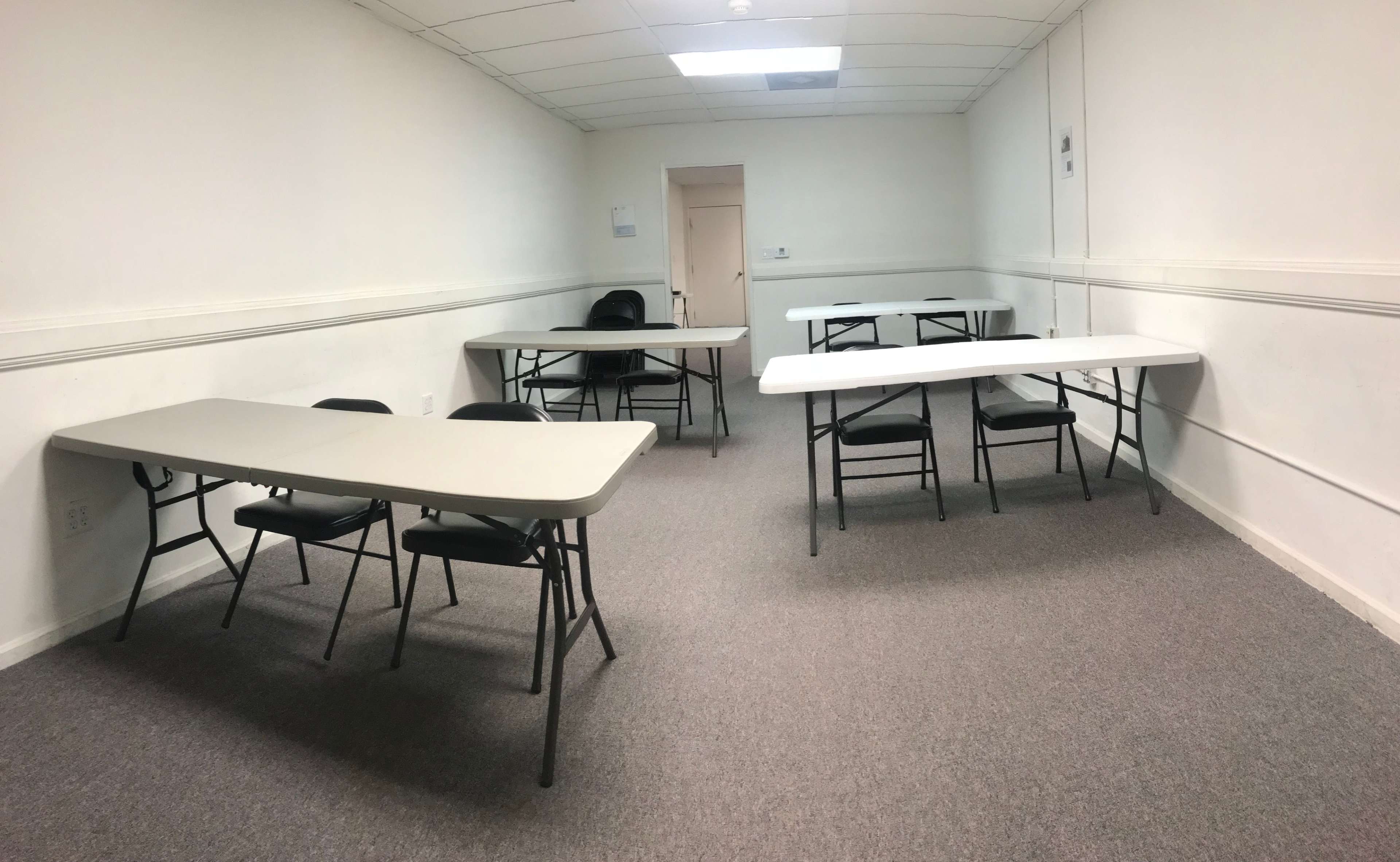 A rectangular room with two sets of folding tables and chairs arranged in a sparse layout against white walls and a beige door.