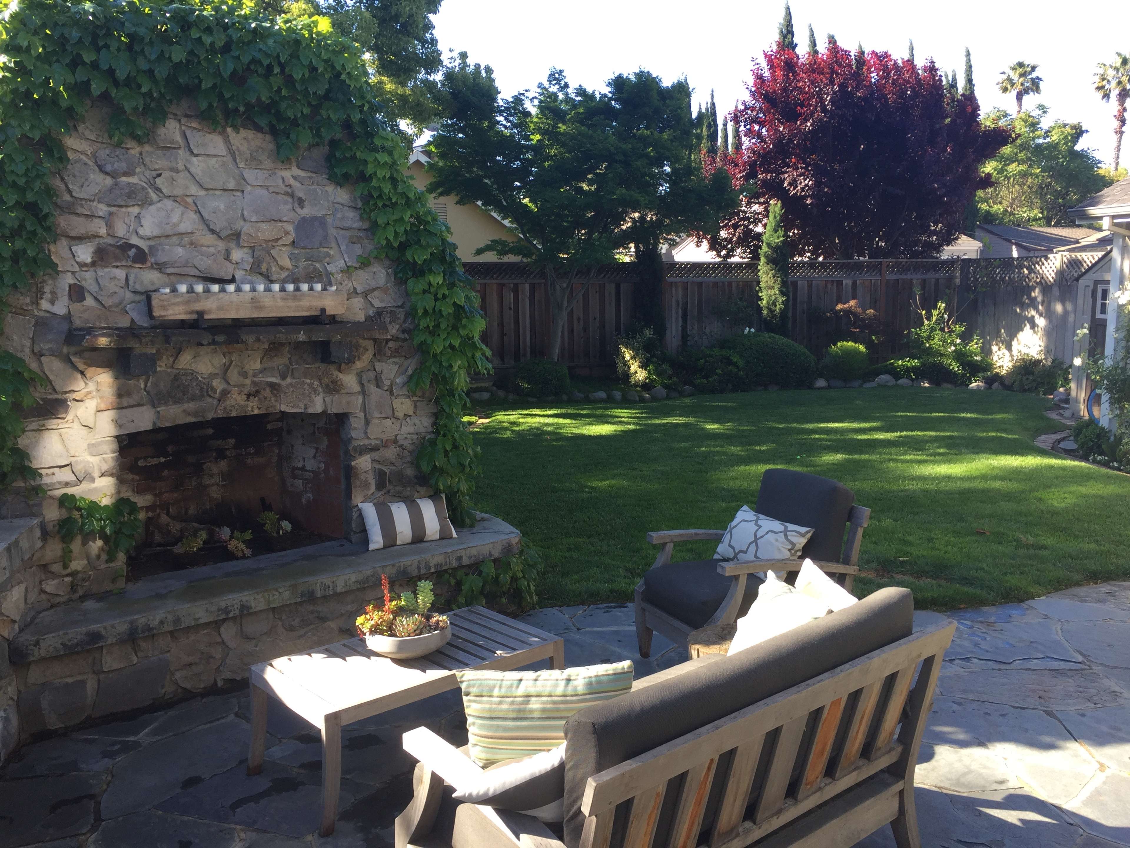 A stone outdoor fireplace is surrounded by a well-maintained lawn and landscaped garden furniture in a residential backyard.