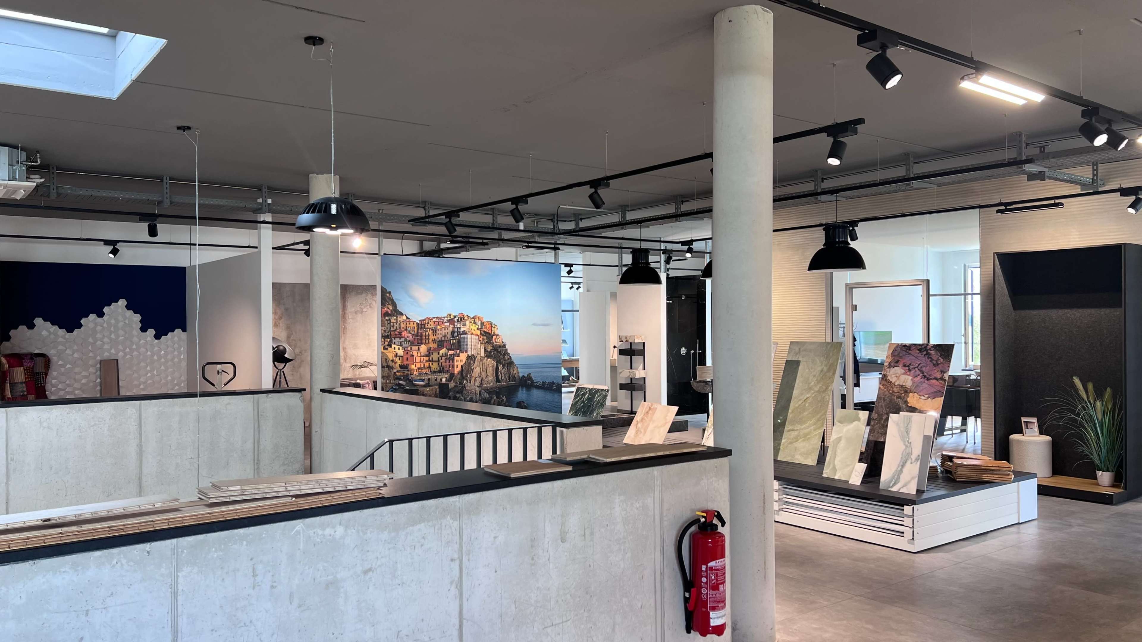 The image shows a modern interior design showroom featuring various display boards and architectural materials, with light fixtures overhead and open areas for showcasing products.