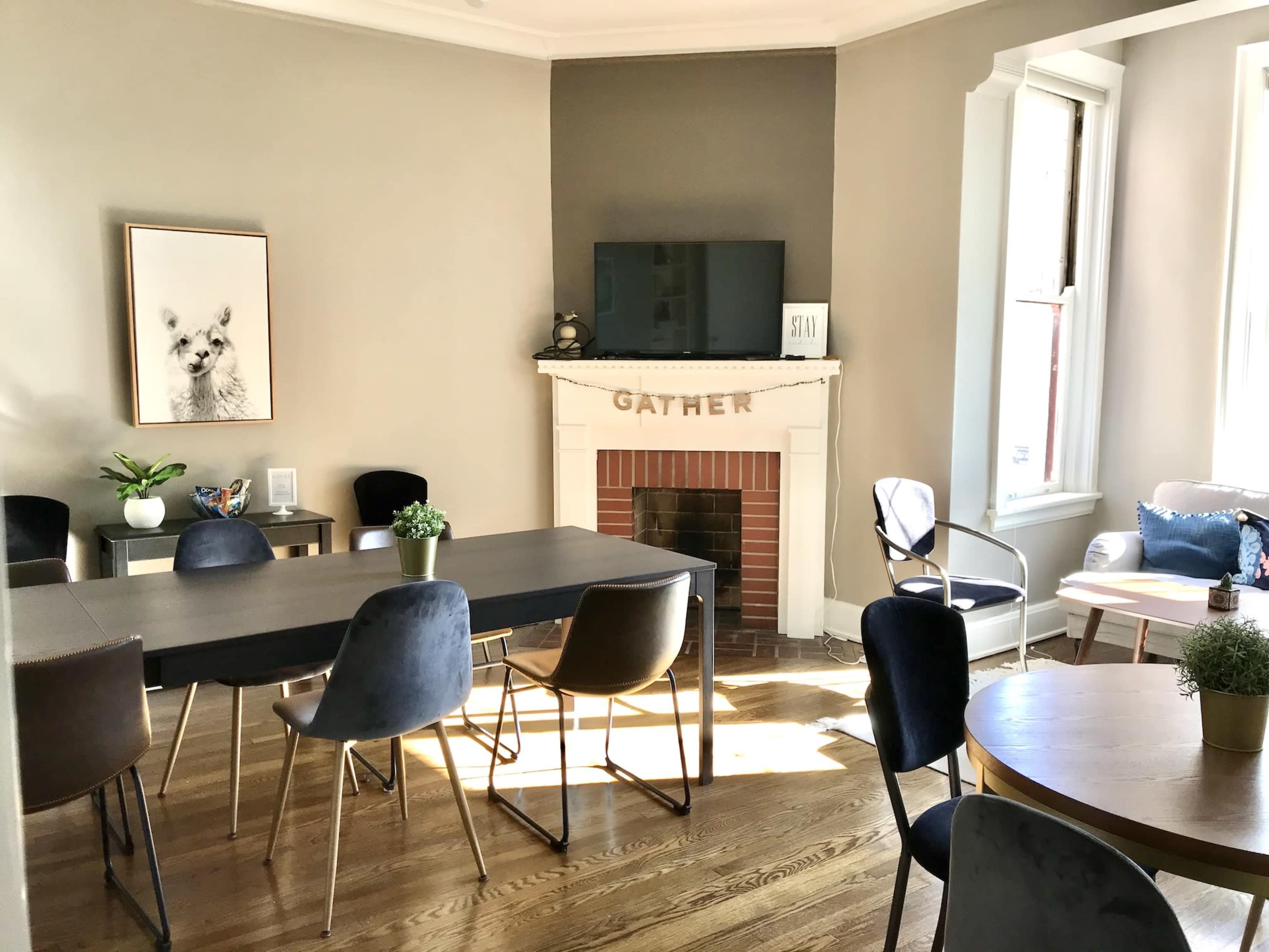 A spacious room features a dining table surrounded by chairs, a television above a fireplace, and various seating areas with plants and artwork on the walls.