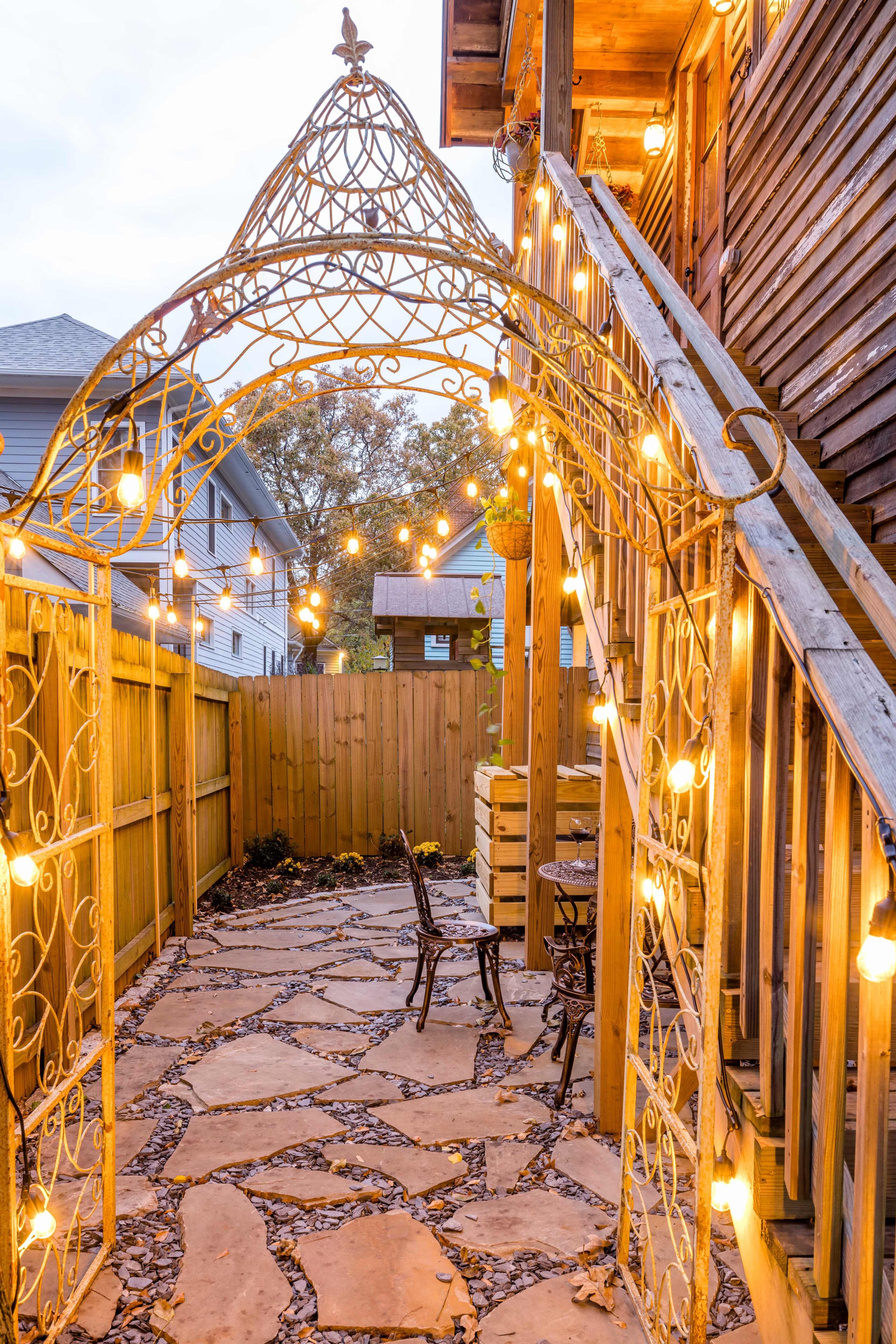 A decorative archway adorned with string lights that leads to a stone pathway, flanked by wooden fences and steps leading to a building.