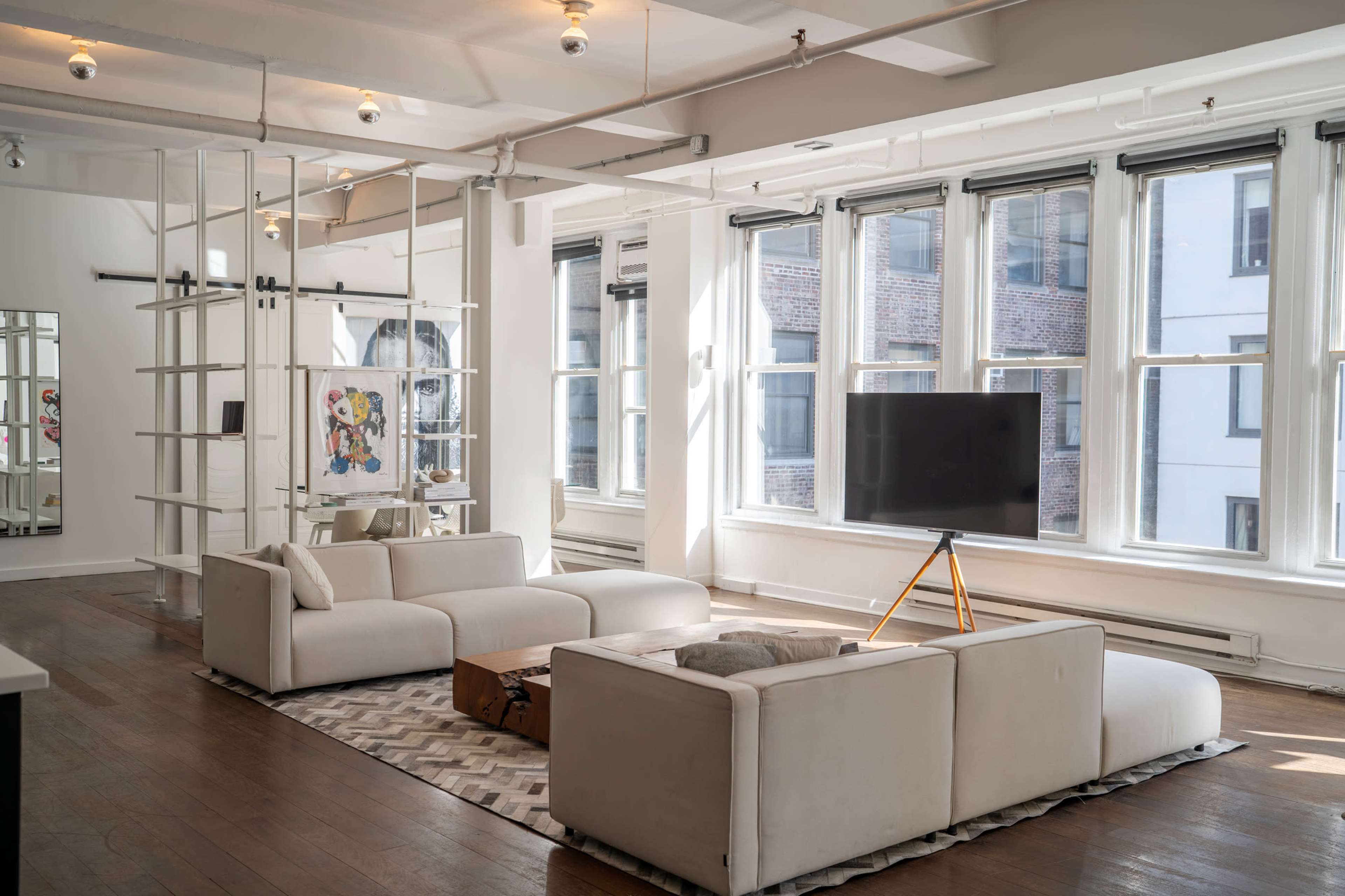 The image shows a modern living room with two white sofas, a wooden coffee table, a TV on a stand, and large windows allowing natural light to fill the space.