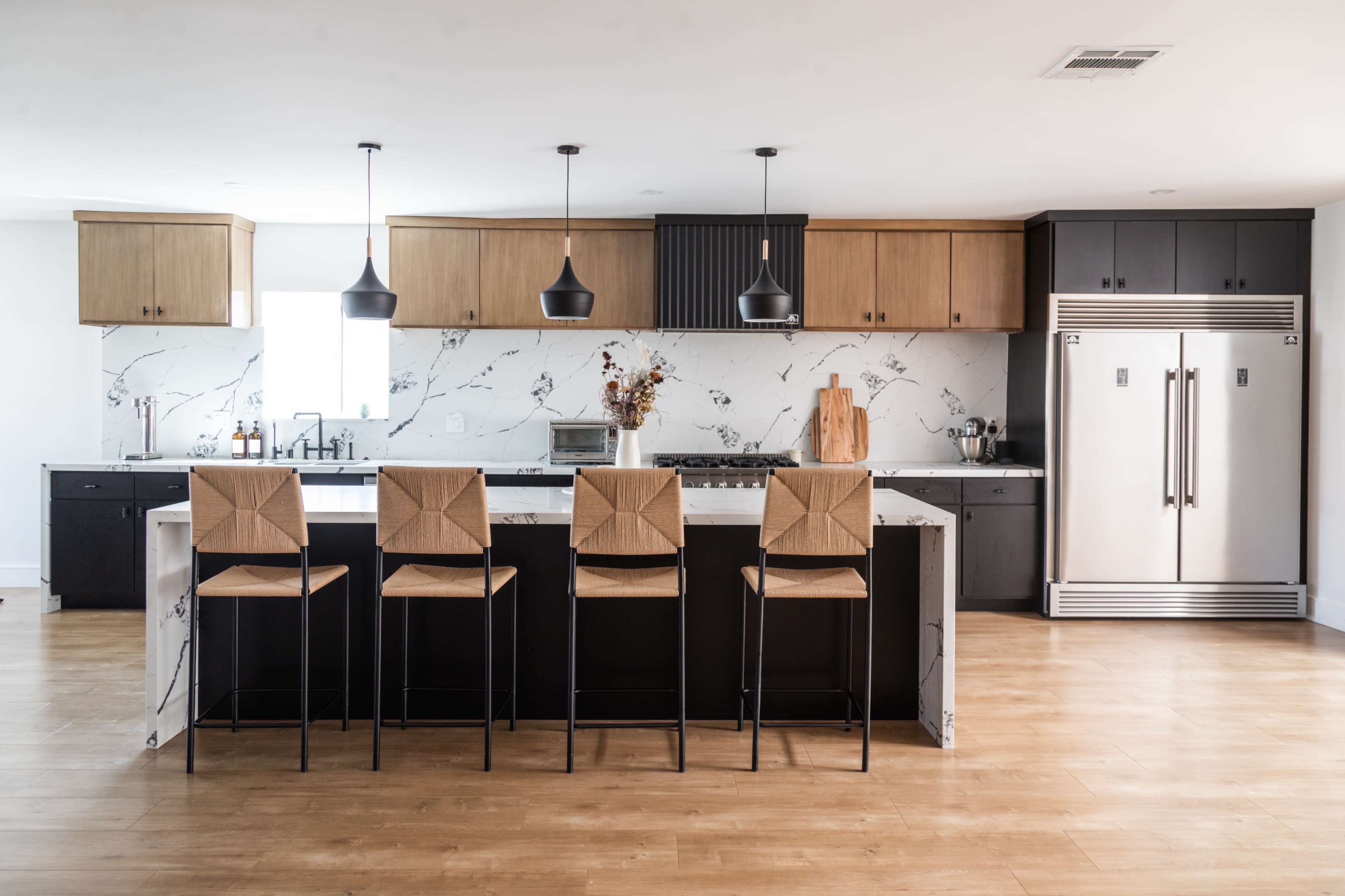 A modern kitchen with a large island, four barstools, and black and wood cabinetry, complemented by marble countertops and stainless steel appliances.