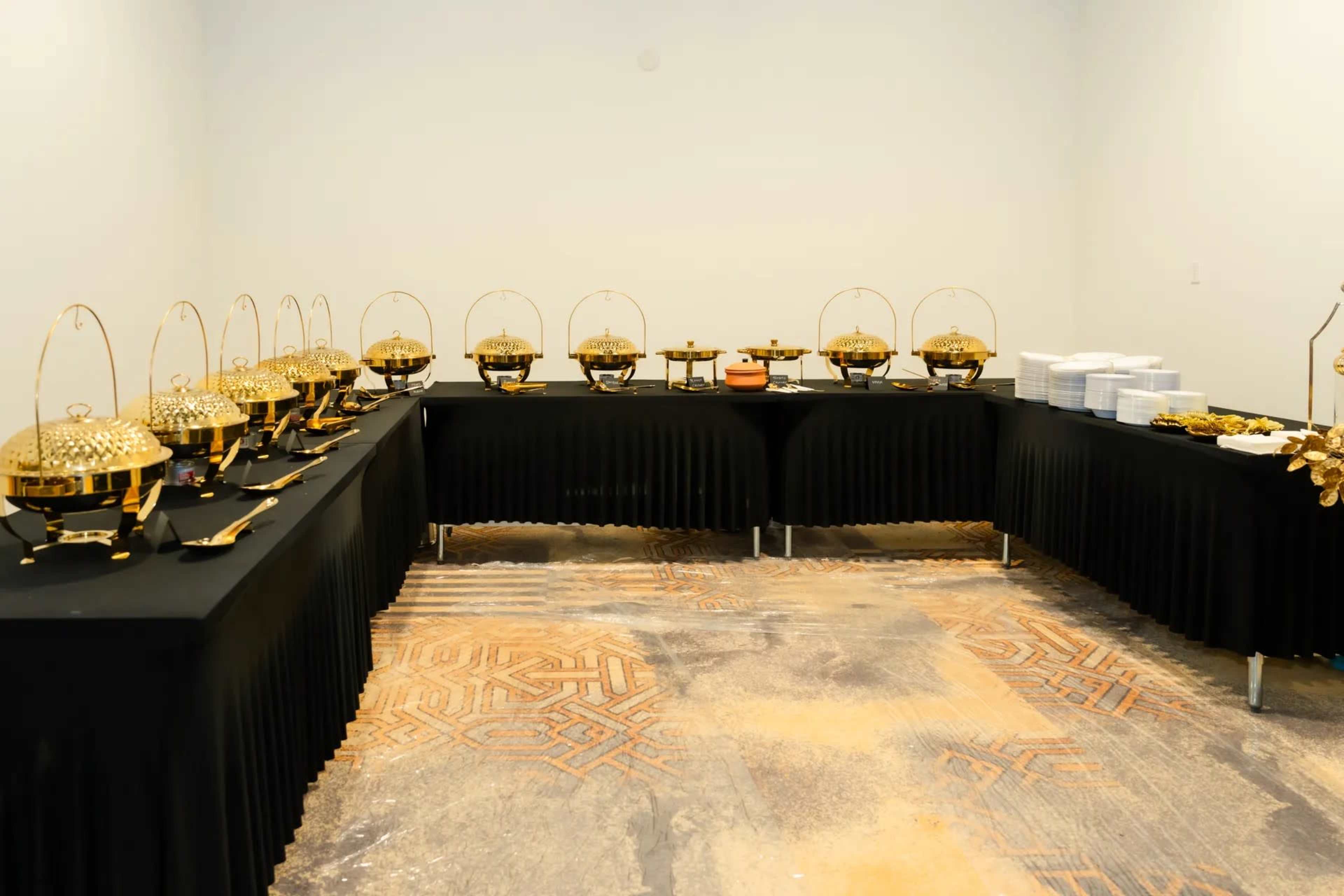A buffet setup features multiple ornate chafing dishes on black tables, accompanied by stacks of white plates.