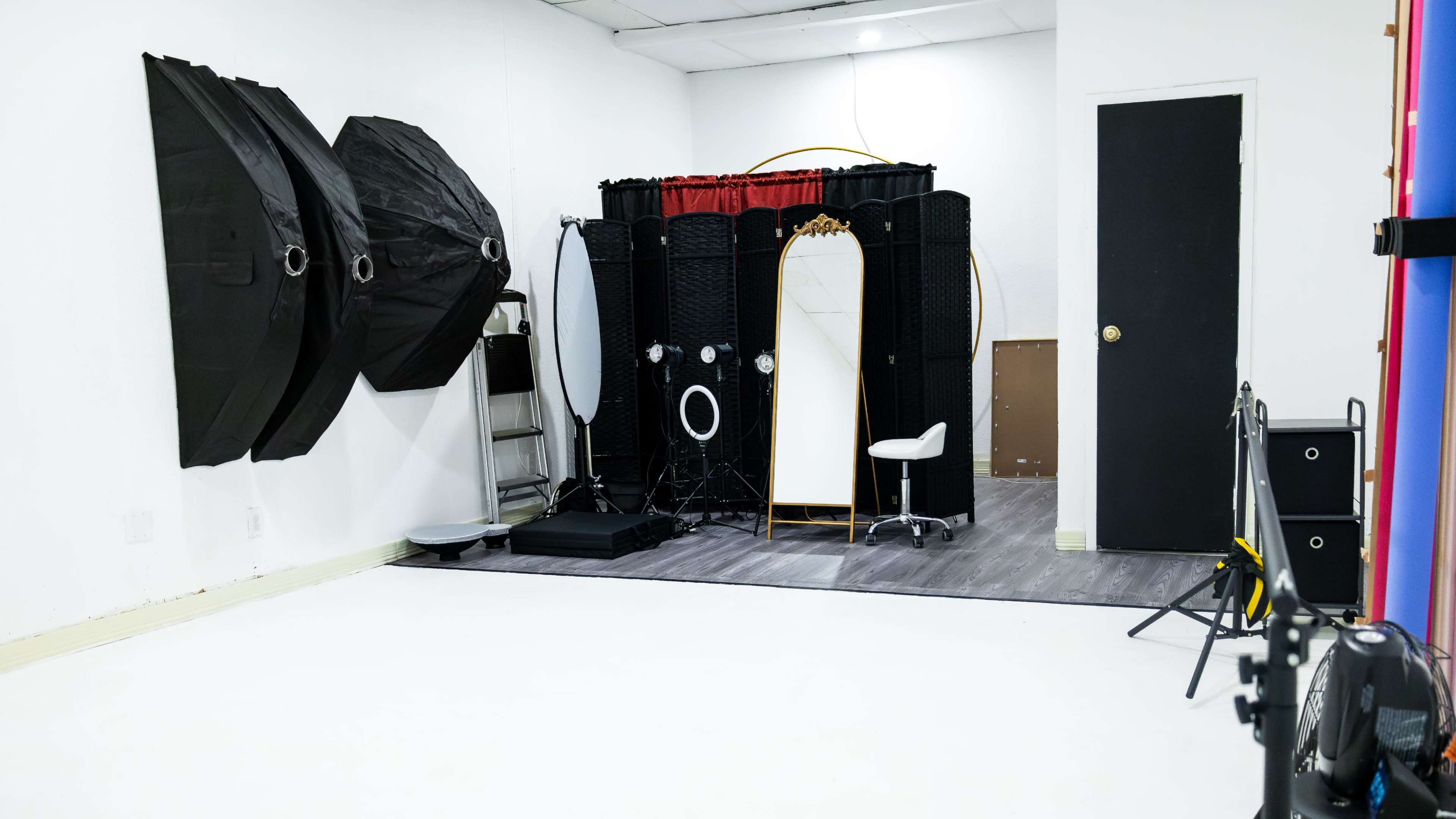 A photography studio featuring black backdrops, a white floor, and a vanity setup with mirrors and lighting equipment.