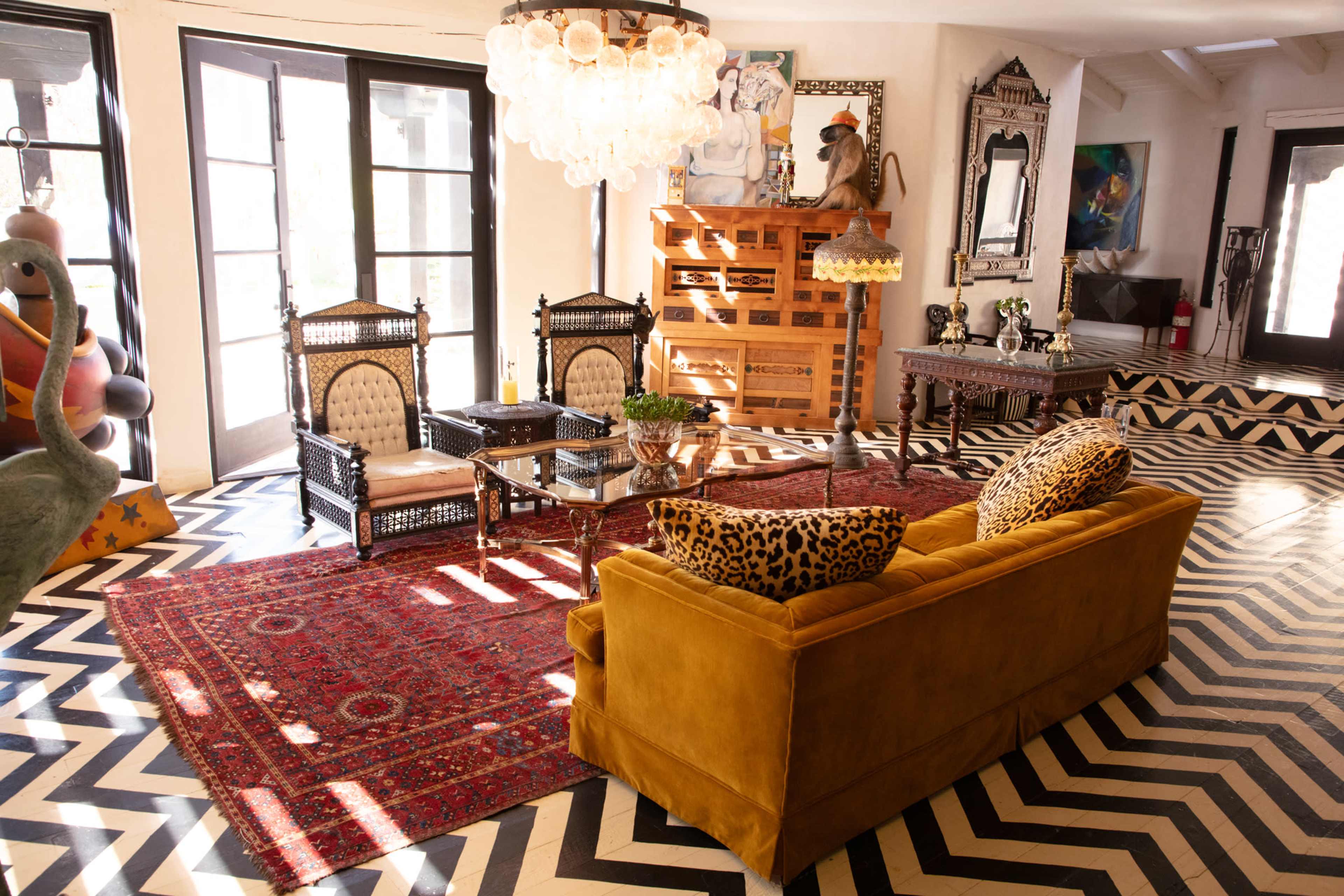 The image shows a stylish living room featuring a yellow sofa, a glass coffee table, decorative chairs, and an intricate rug on a black and white chevron floor.