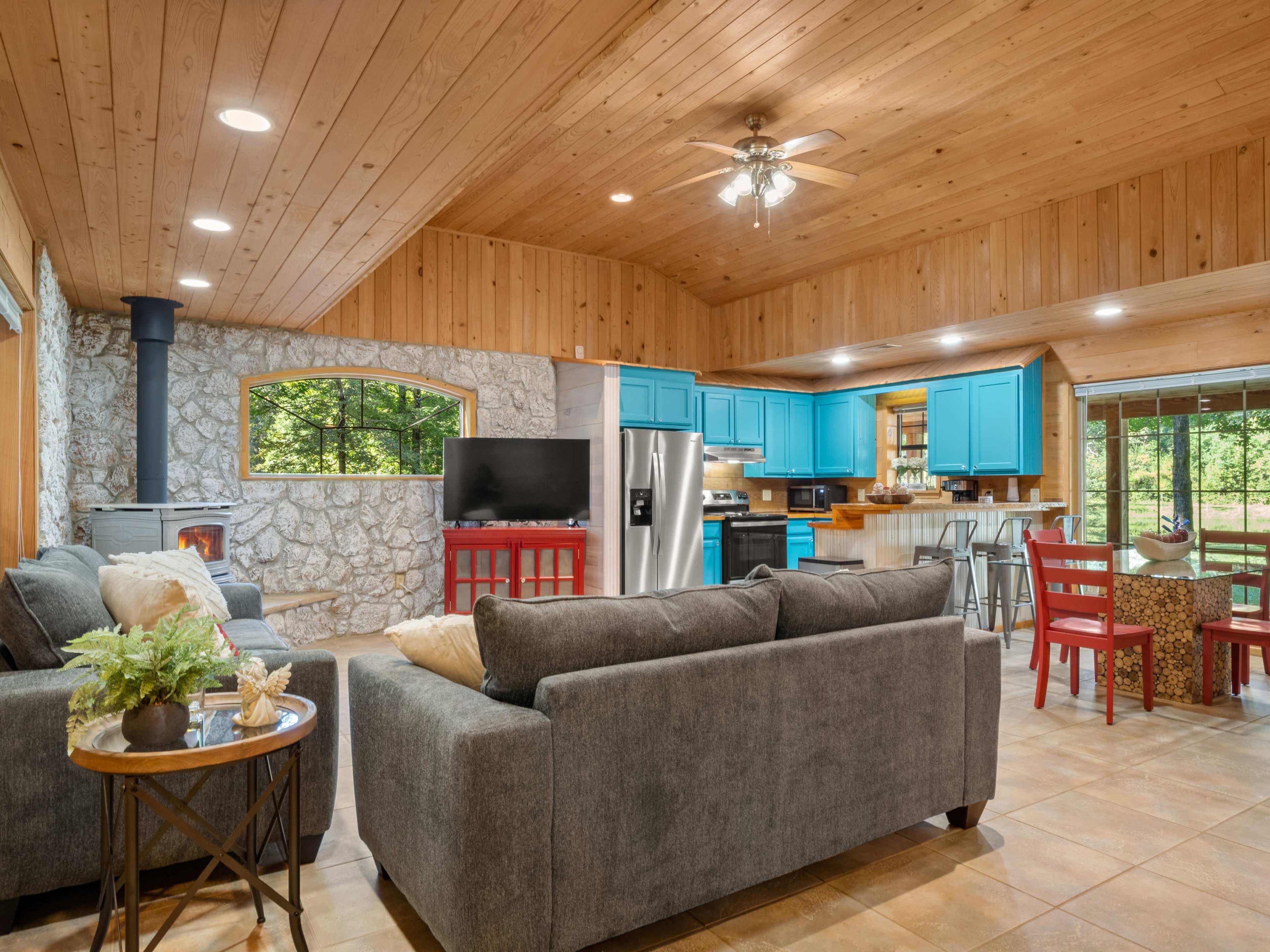 A rustic living space with wooden ceilings, a stone fireplace, and modern blue cabinetry, featuring a cozy arrangement of furniture and large windows that overlook a natural setting.