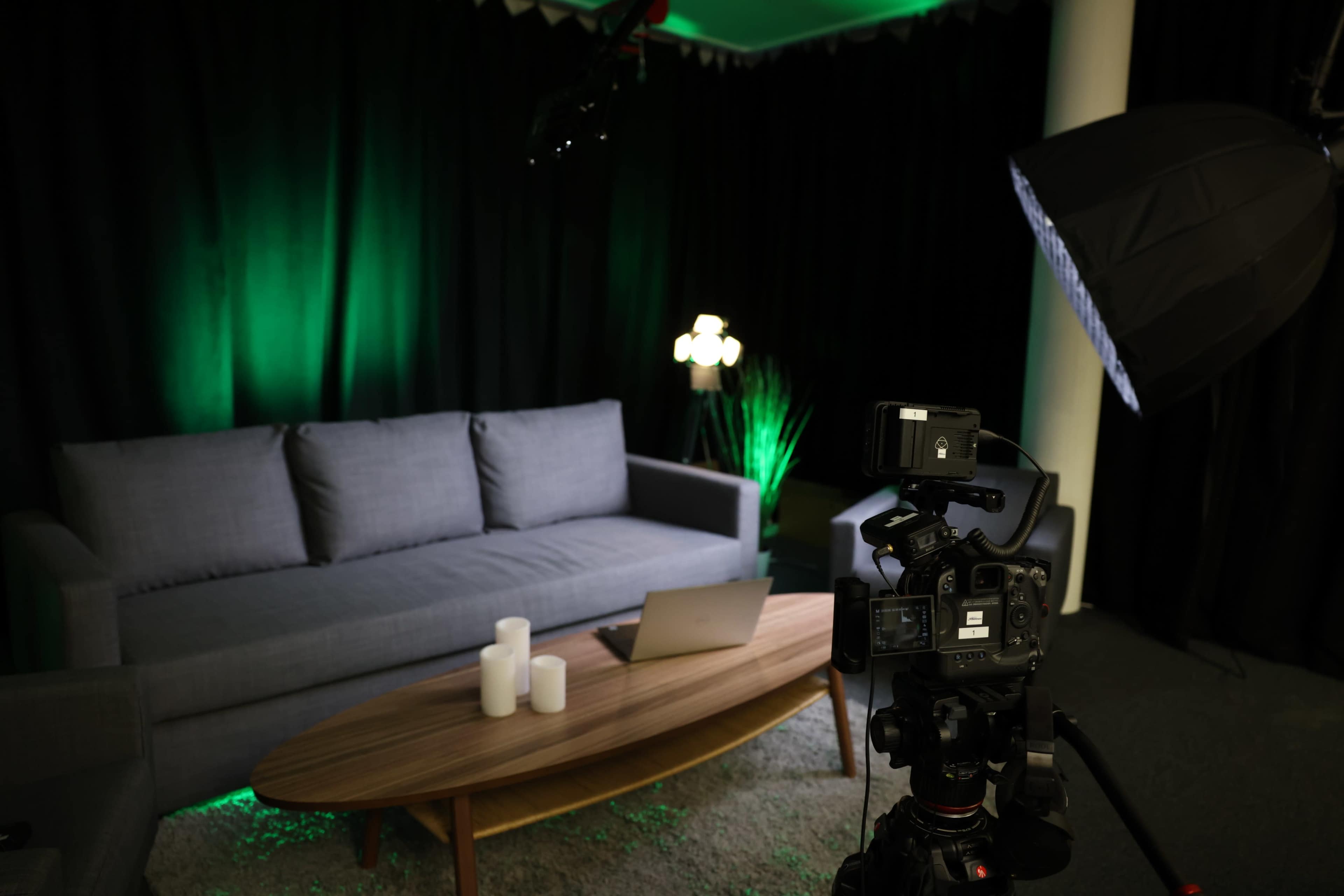 A modern living room setup features a gray sofa, a coffee table with candles and a laptop, and is illuminated by green lights against a black backdrop.