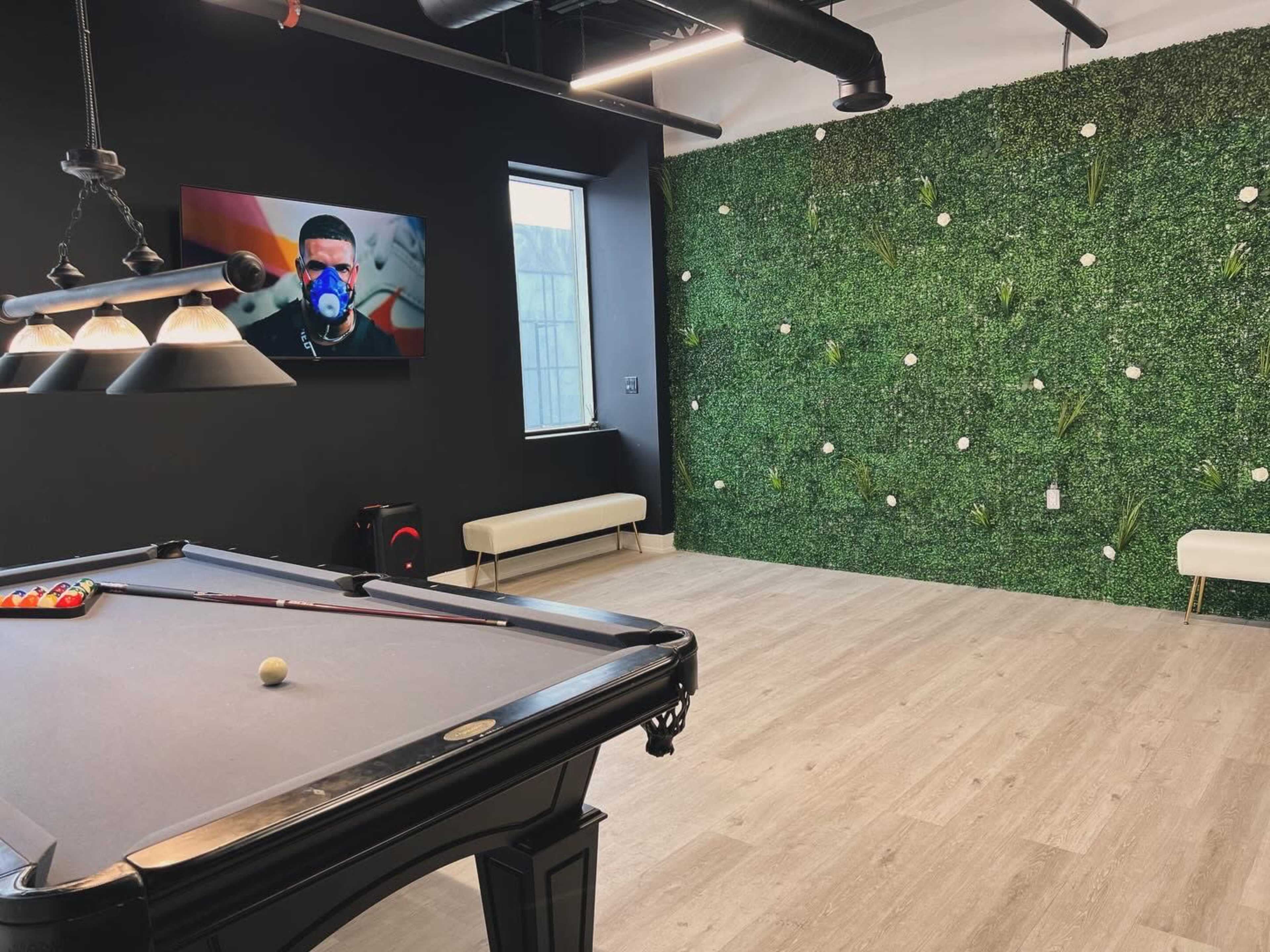 The scene features a pool table in a room with a green wall of artificial plants and a television displaying a person wearing a mask.