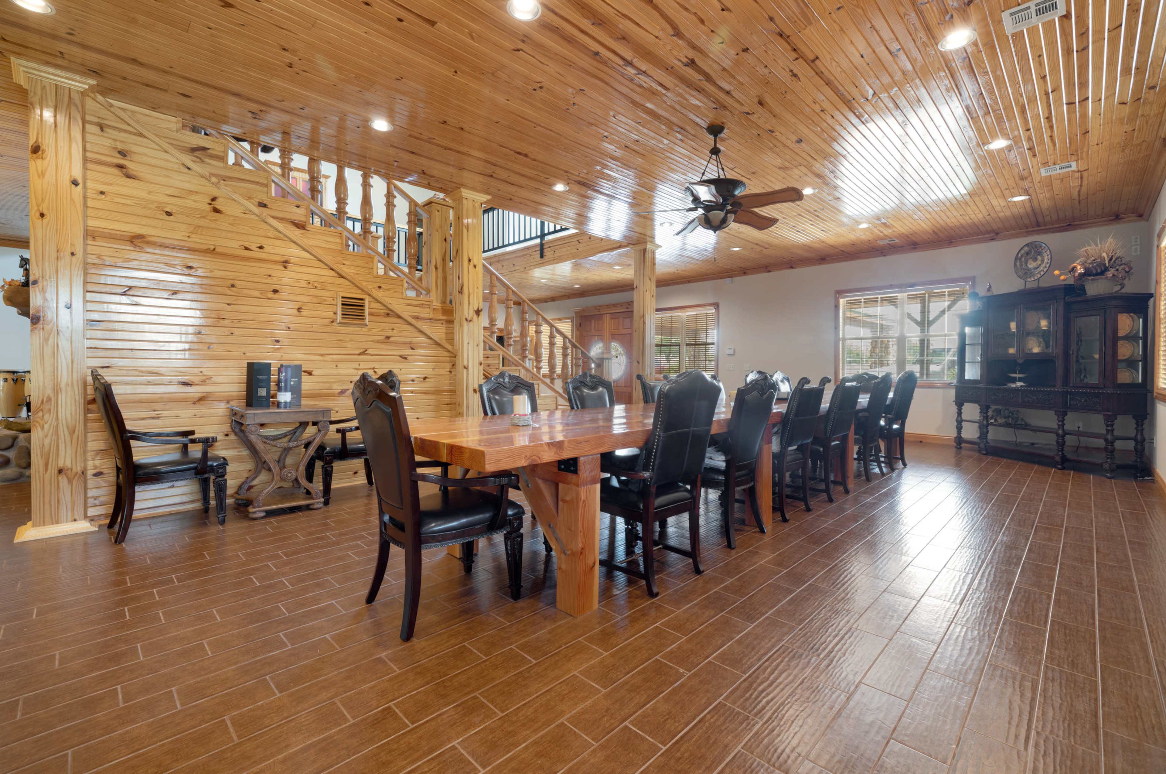 The room features a wooden dining table surrounded by black chairs, with polished wooden walls and ceiling, adjacent to a staircase and a large window.