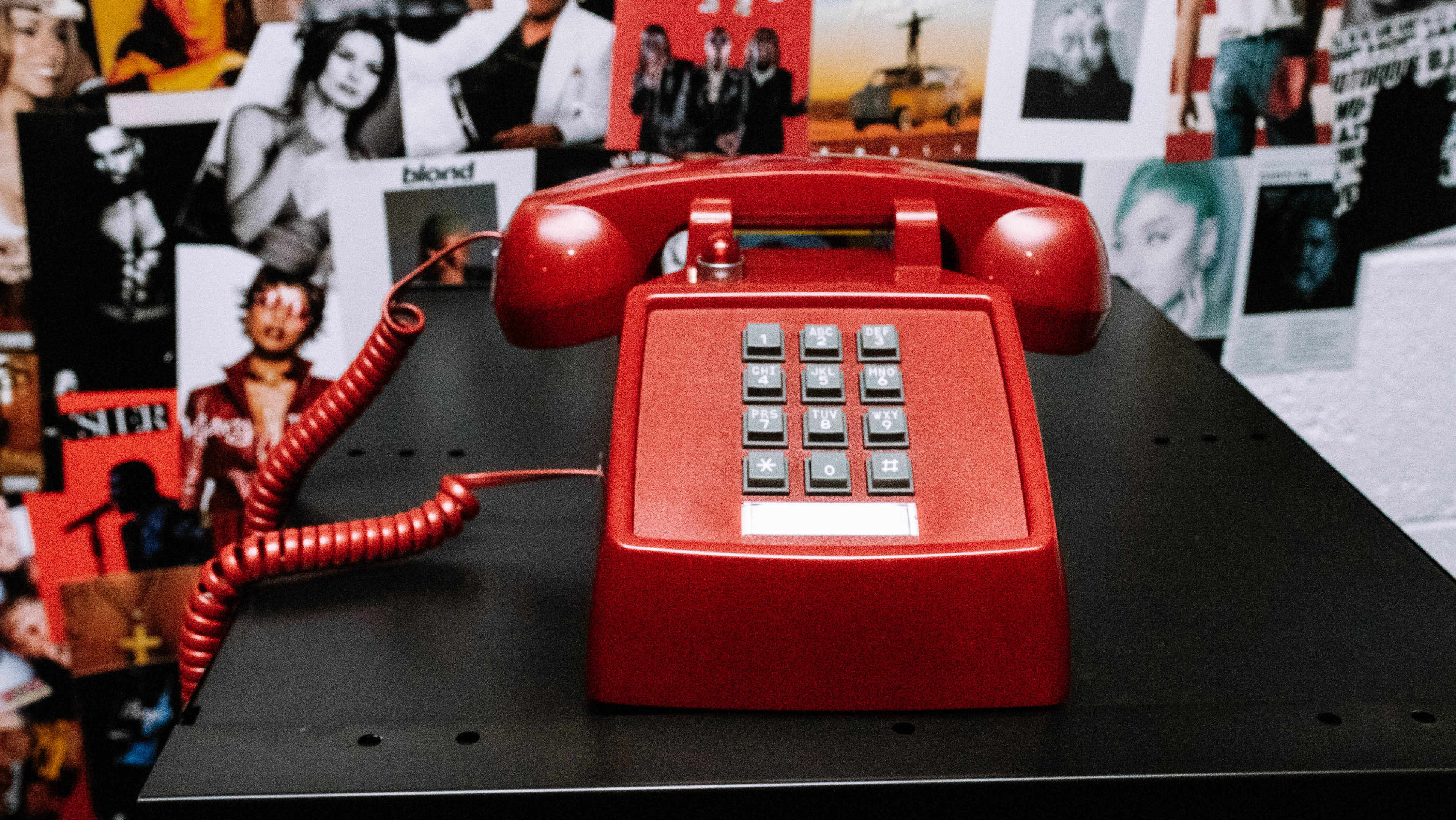 A bright red vintage telephone sits on a black surface in front of a wall covered with various photographs.