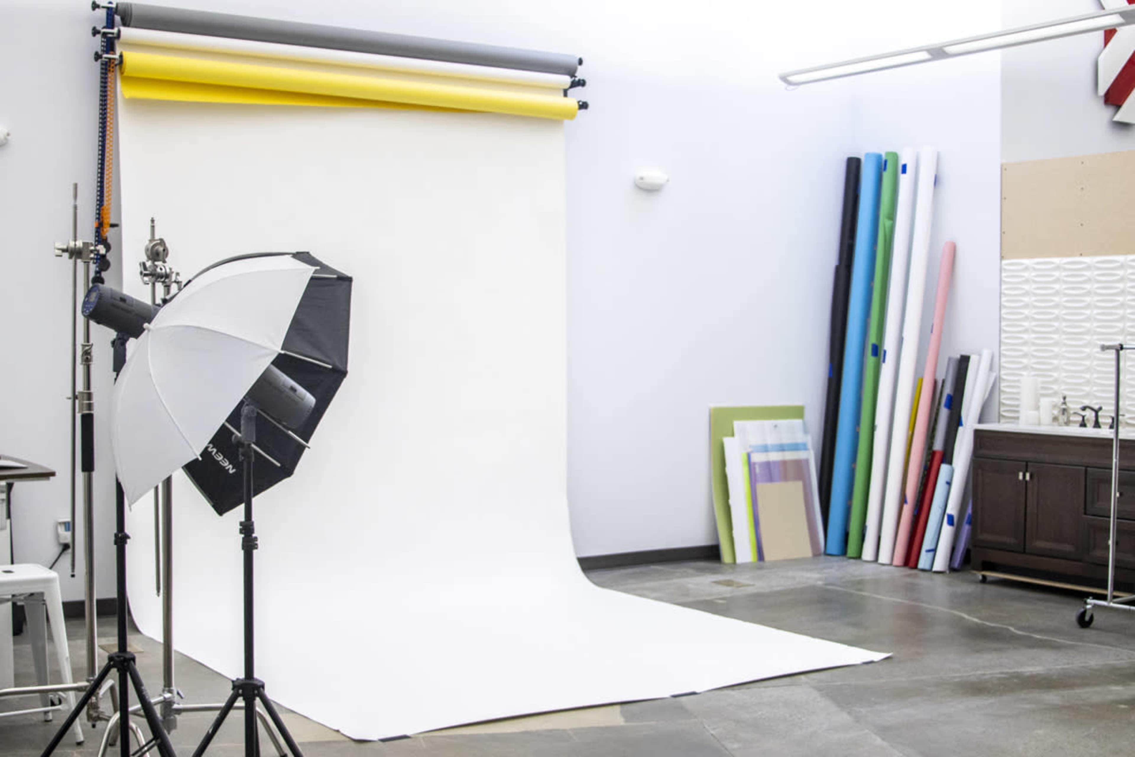 A photography studio is set up with a white backdrop, lighting equipment, and rolls of colored paper stacked against the wall.