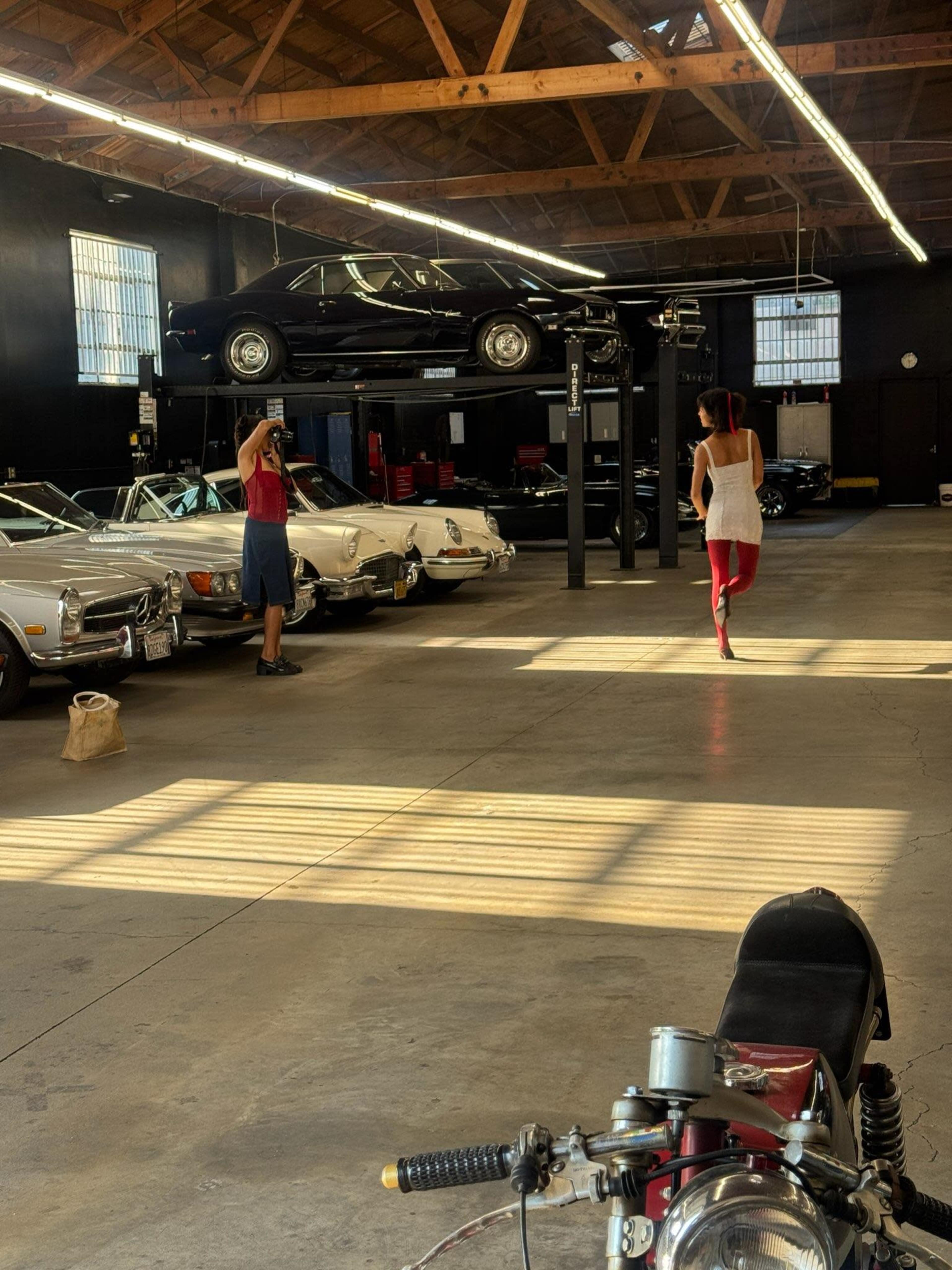A man and a woman are in a garages displaying classic cars, with a black car elevated on a lift in the background.