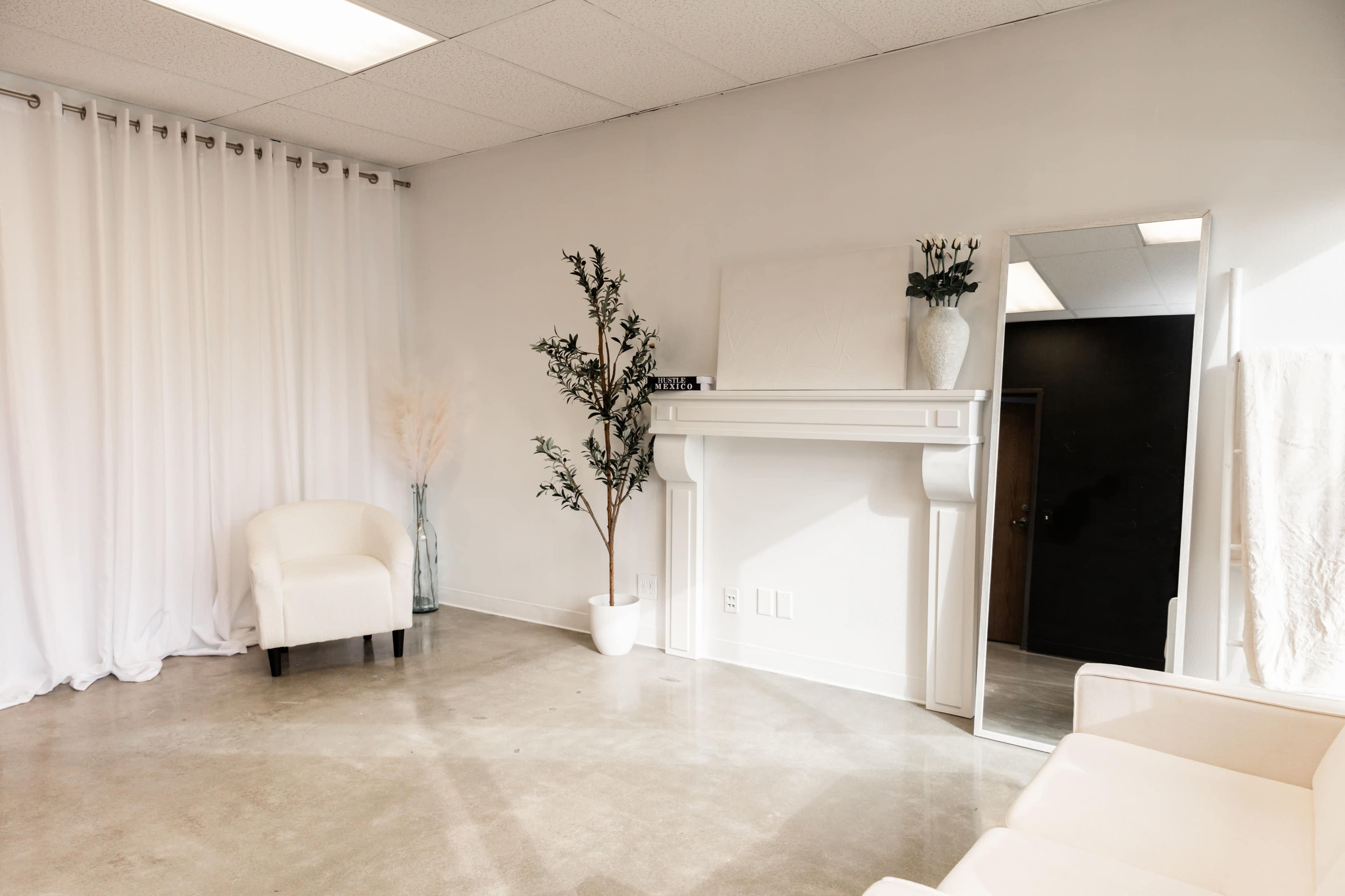 A minimalist room featuring a white wall, a decorative fireplace with a vase, a potted plant, a full-length mirror, and two light-colored chairs.