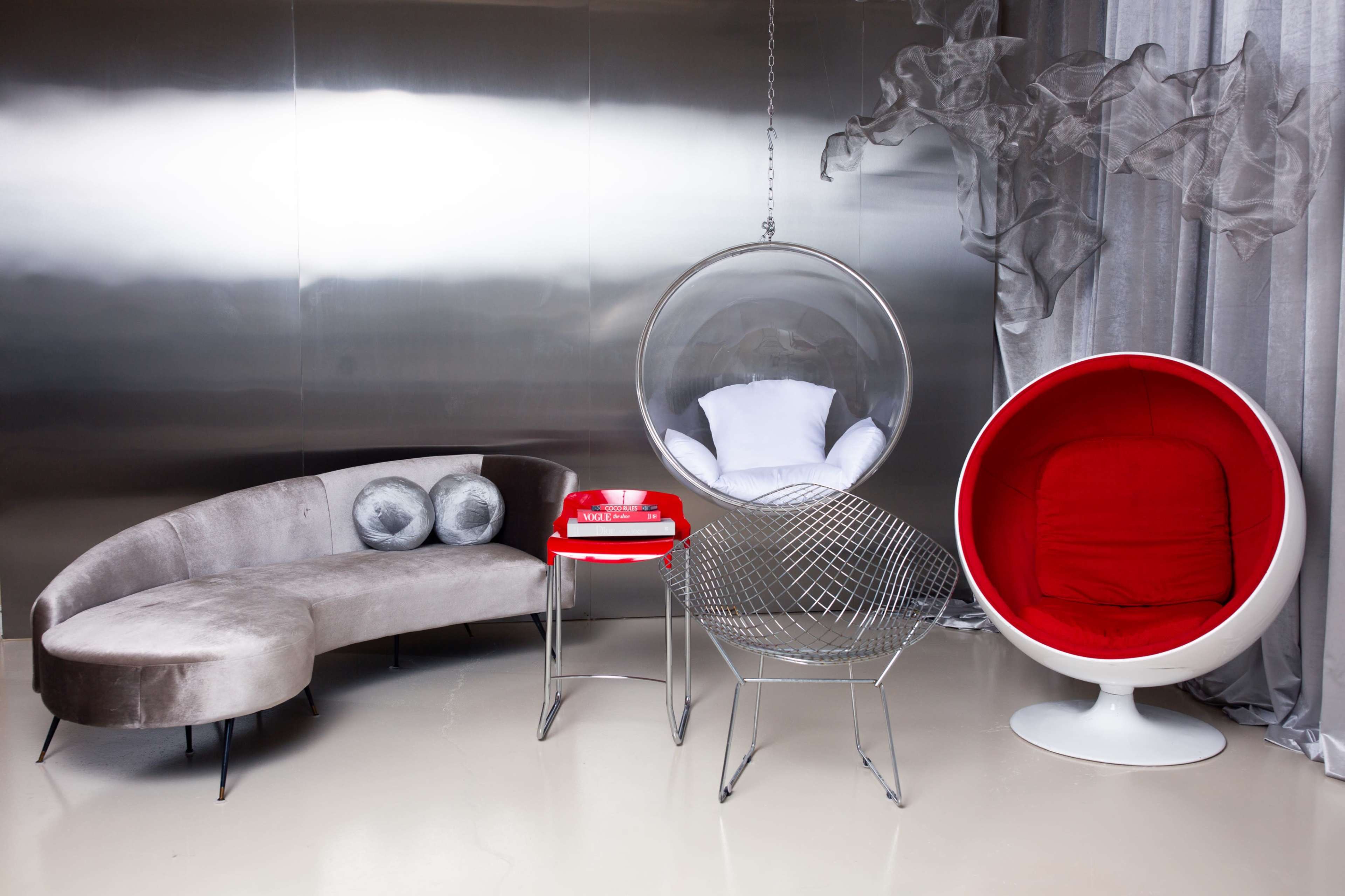 The image features a modern interior setting with a curved gray sofa, a round hanging chair, a wireframe chair, and a circular red chair.