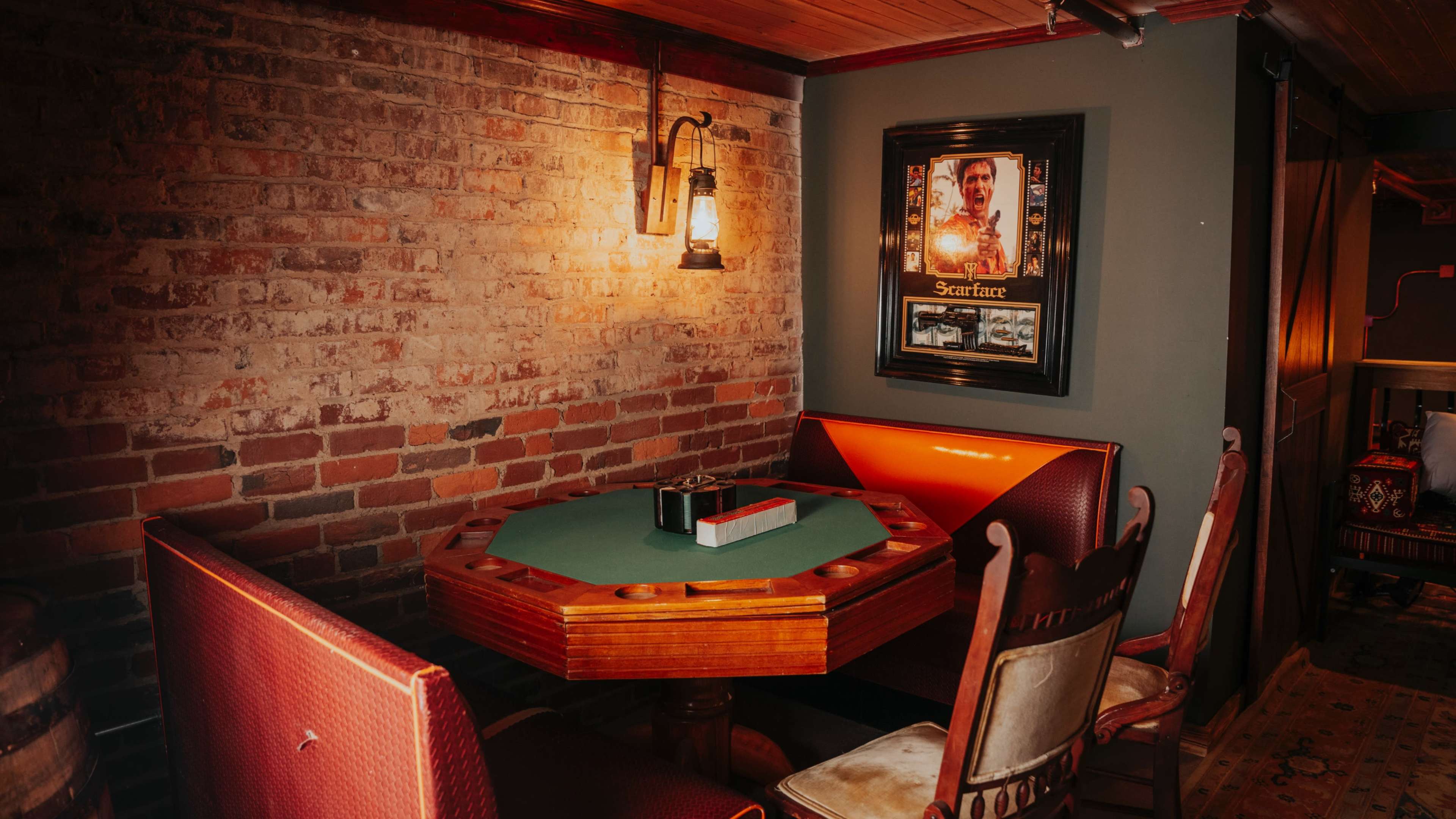 A small poker table with a green felt surface is surrounded by wooden chairs in a dimly lit room with exposed brick walls and a framed poster on the wall.
