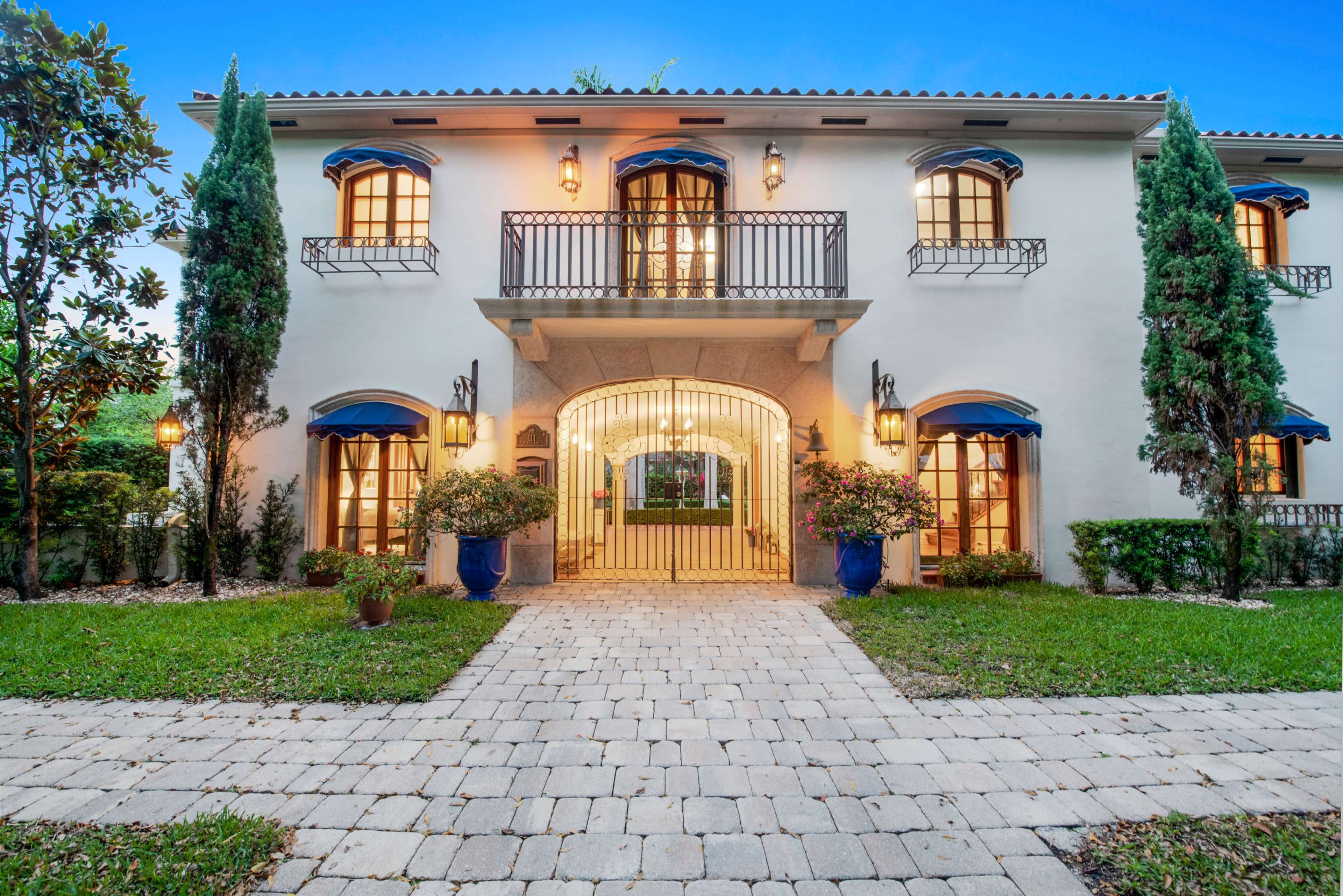 A two-story, Mediterranean-style house with blue awnings and a decorative entrance is surrounded by well-maintained greenery and paving stones.